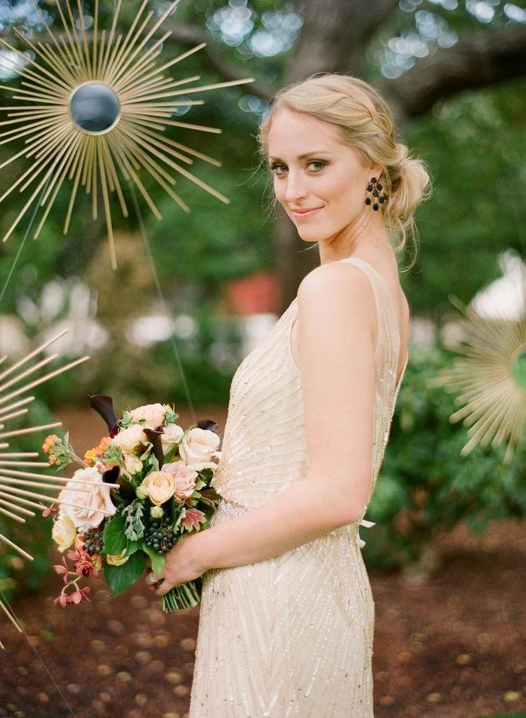 Young woman in a cream-colored dress holding a bouquet of pink, cream, and dark purple flowers, standing outdoors with trees and decorative golden starburst ornaments around her.
