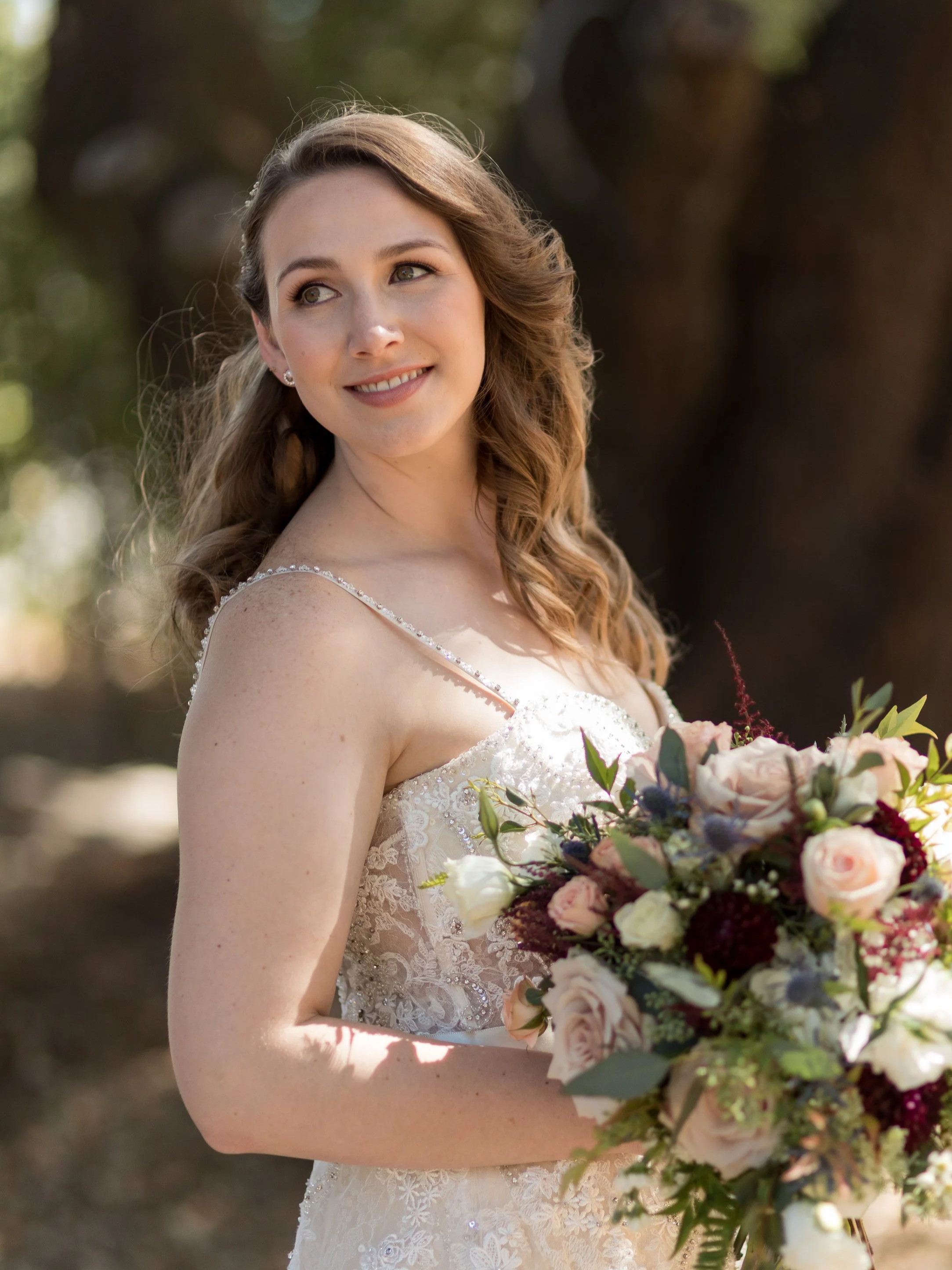 A smiling bride in a white lace wedding dress holding a bouquet of roses and greenery outdoors.