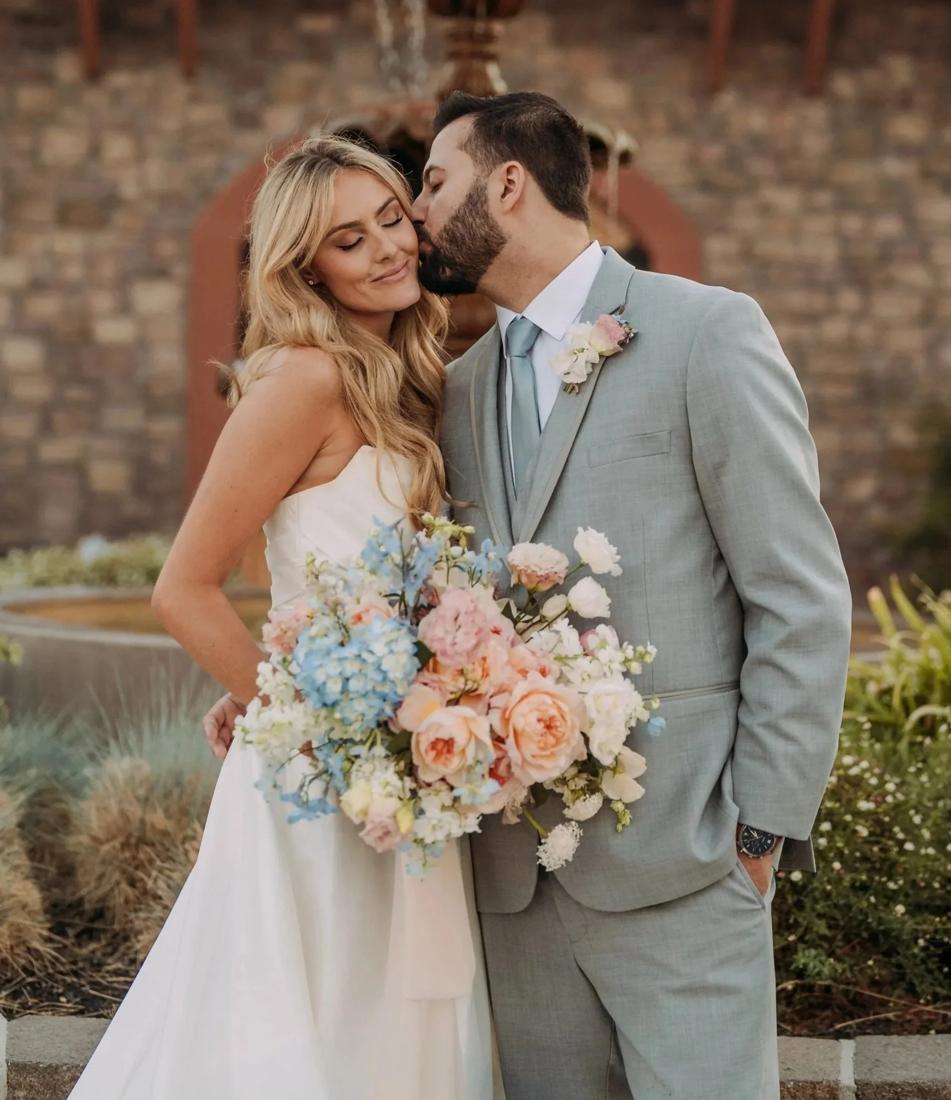 A couple dressed in wedding attire, with the woman holding a large bouquet of pastel-colored flowers, sharing a moment close to each other outdoors against a brick wall background.
