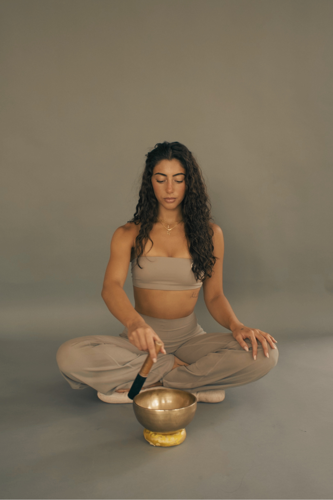 A woman with long curly hair sitting cross-legged on the floor, dressed in beige athletic wear, performing a meditation with a singing bowl in front of her, holding a mallet, against a plain grey background.