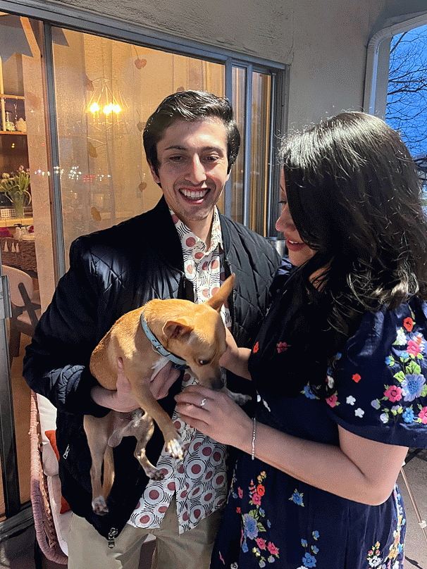 A man and woman looking at a small tan dog the woman is holding. They are standing near a glass door outside in an evening setting.