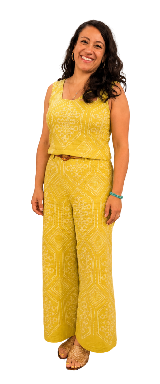 A woman with long, dark, wavy hair smiling and wearing a yellow patterned sleeveless jumpsuit, accessorized with gold hoop earrings, a necklace, a turquoise bracelet, and open-toed shoes.