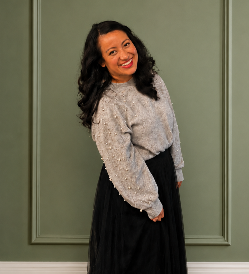 Smiling woman with long, dark, curly hair wearing a gray sweater with pearl embellishments and a black skirt, standing in front of a green wall.