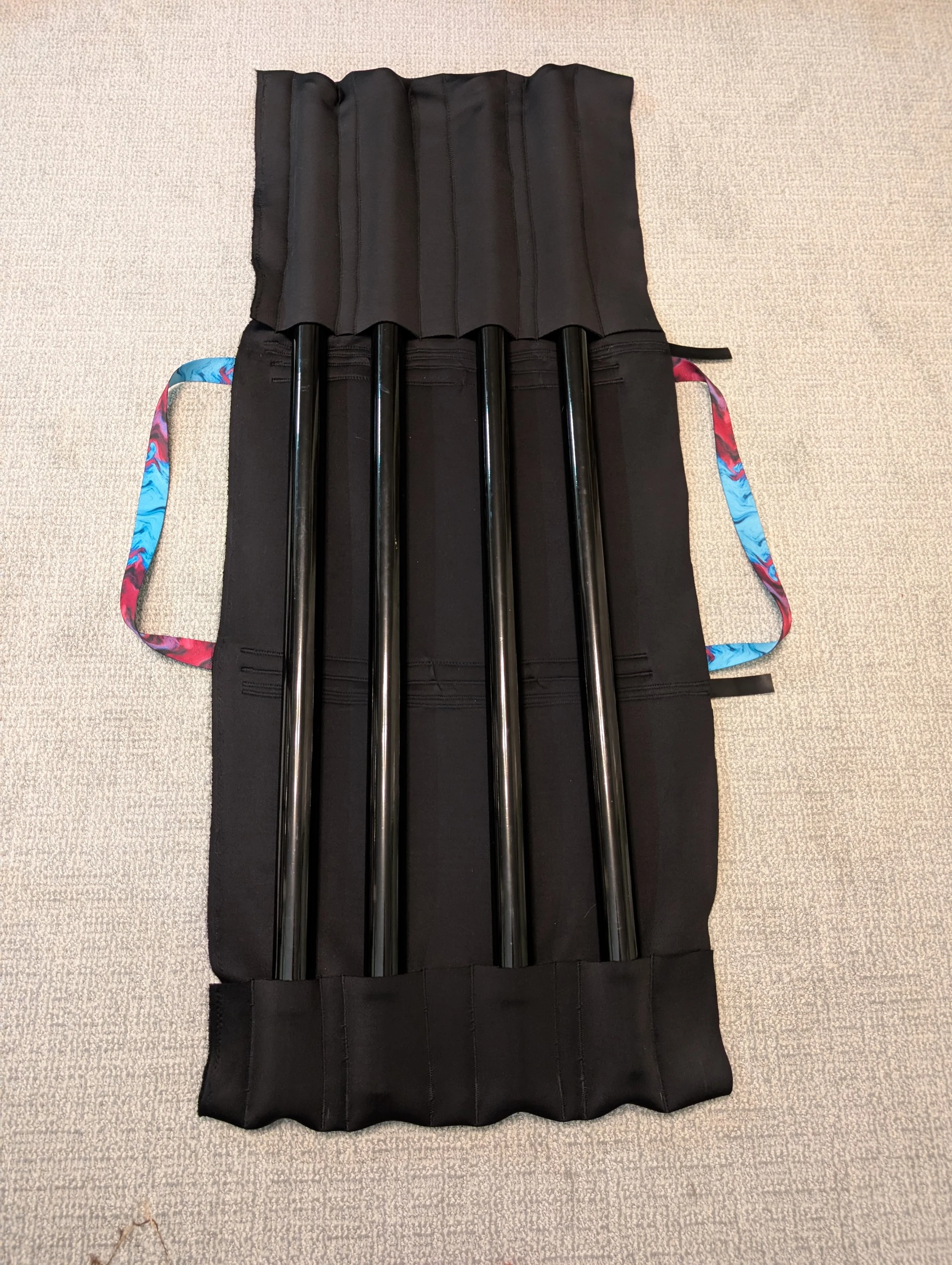 A black storage organizer with four vertical compartments holding four black poles, laid out on a black fabric surface with fabric handles on both sides, on a light beige carpet.