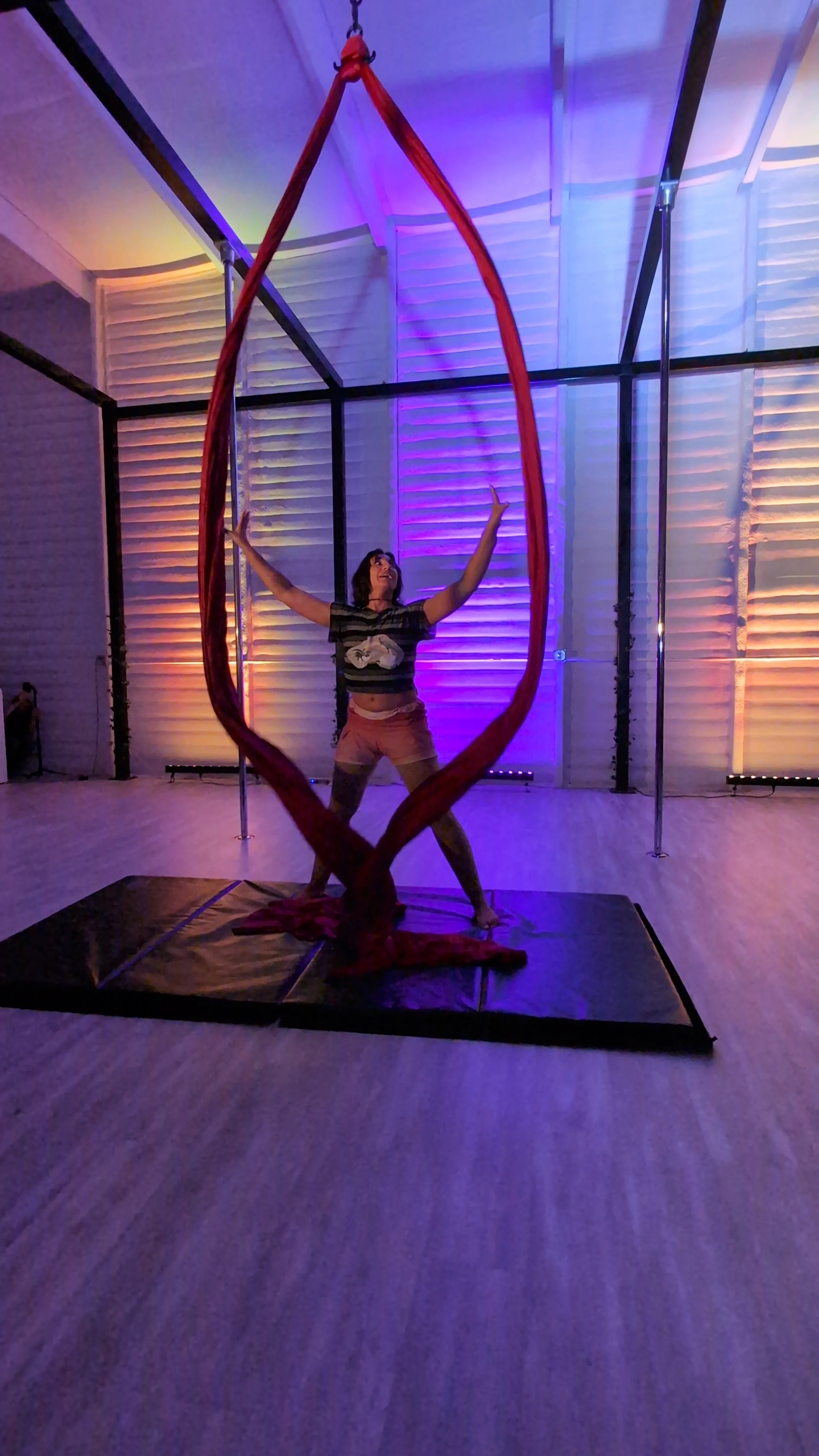 A woman performing an aerial silk routine inside a room illuminated with purple and yellow lights. She is holding onto red fabric with her arms extended and legs apart, standing on a black padded mat.
