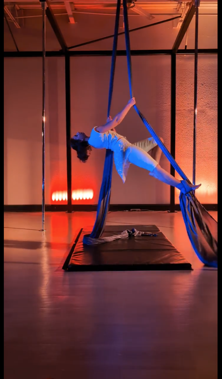 A woman performing aerial silk acrobatics on a stage with a padded mat beneath her, illuminated by warm colorful lighting.