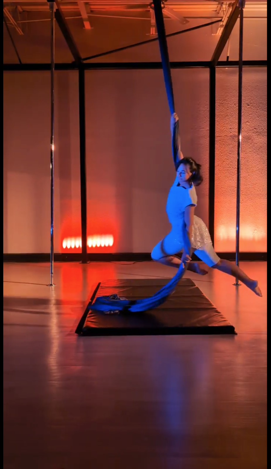 A woman performs aerial silk acrobatics on a black fabric in a dimly lit room with red and orange lighting.
