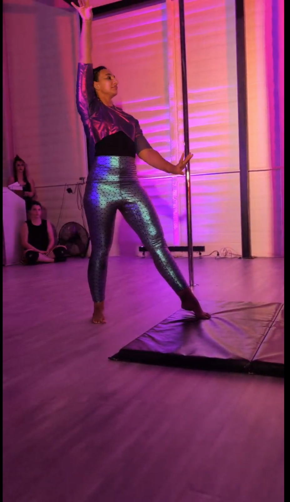 A woman posing indoors with purple lighting, wearing shiny, mermaid patterned leggings and a purple top.