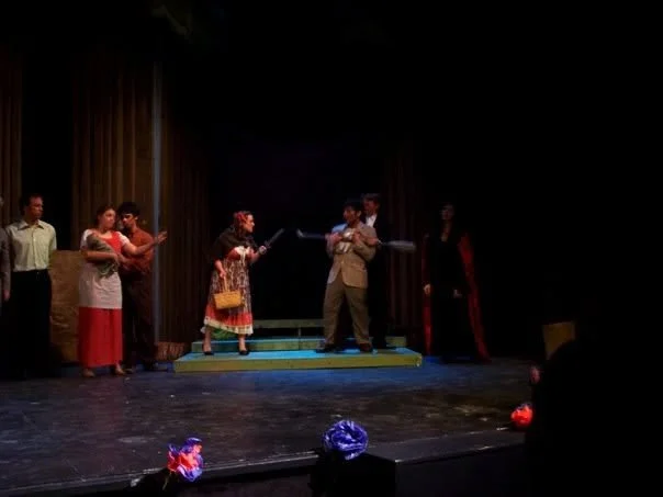 A theatrical stage with six actors in costume, some holding scripts, with props and set pieces, under stage lights.