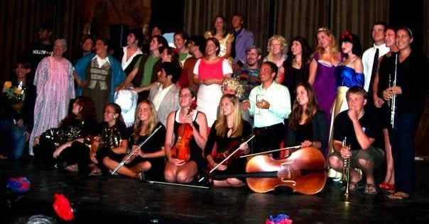 Group of performers, some holding musical instruments, dressed in colorful costumes, standing and sitting on stage with a dark backdrop.