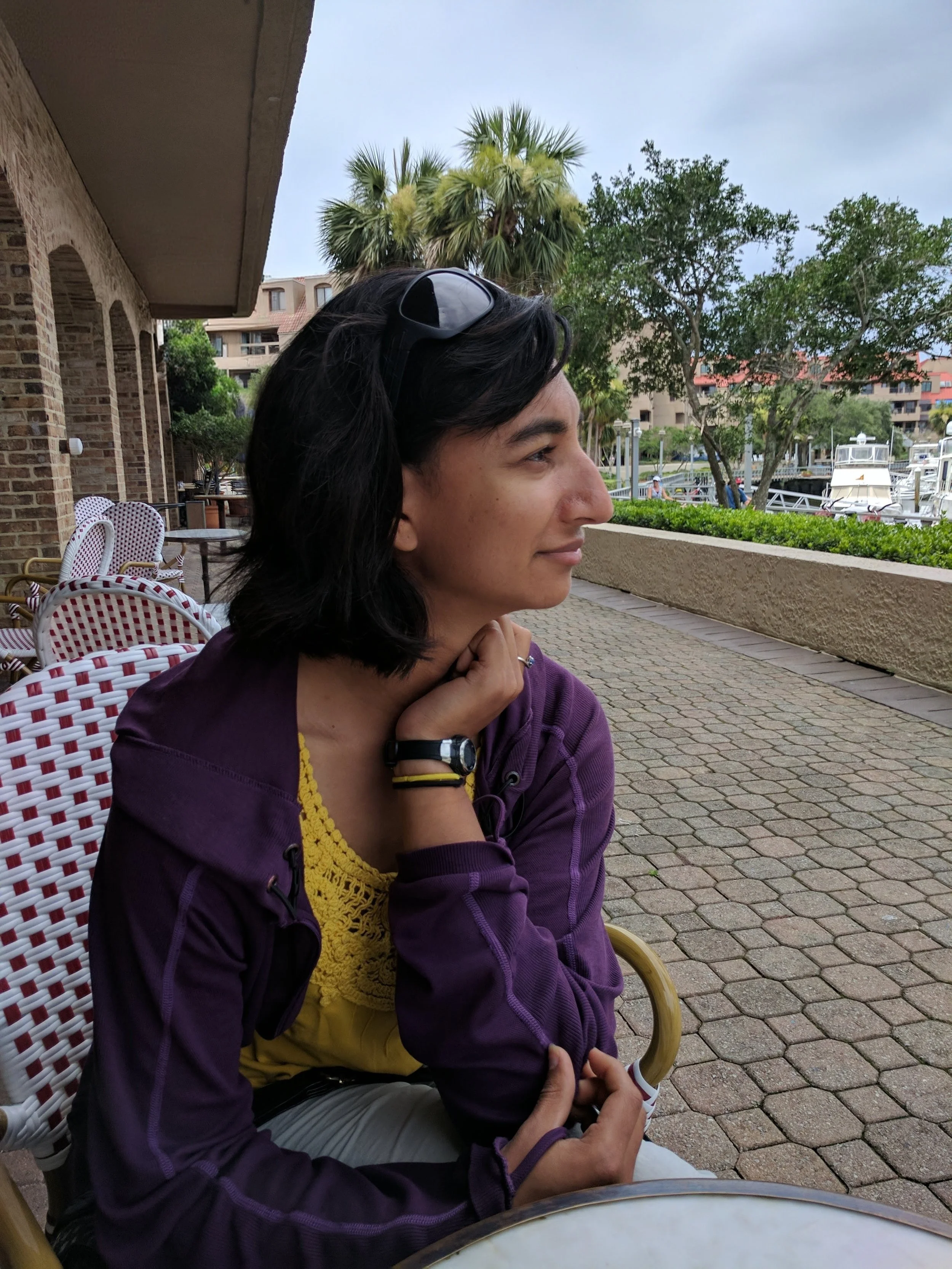 Side profile of a woman sitting outdoors at a cafe, wearing a purple jacket and yellow top, with sunglasses on her head, with palm trees, a tree, and boats in the background.