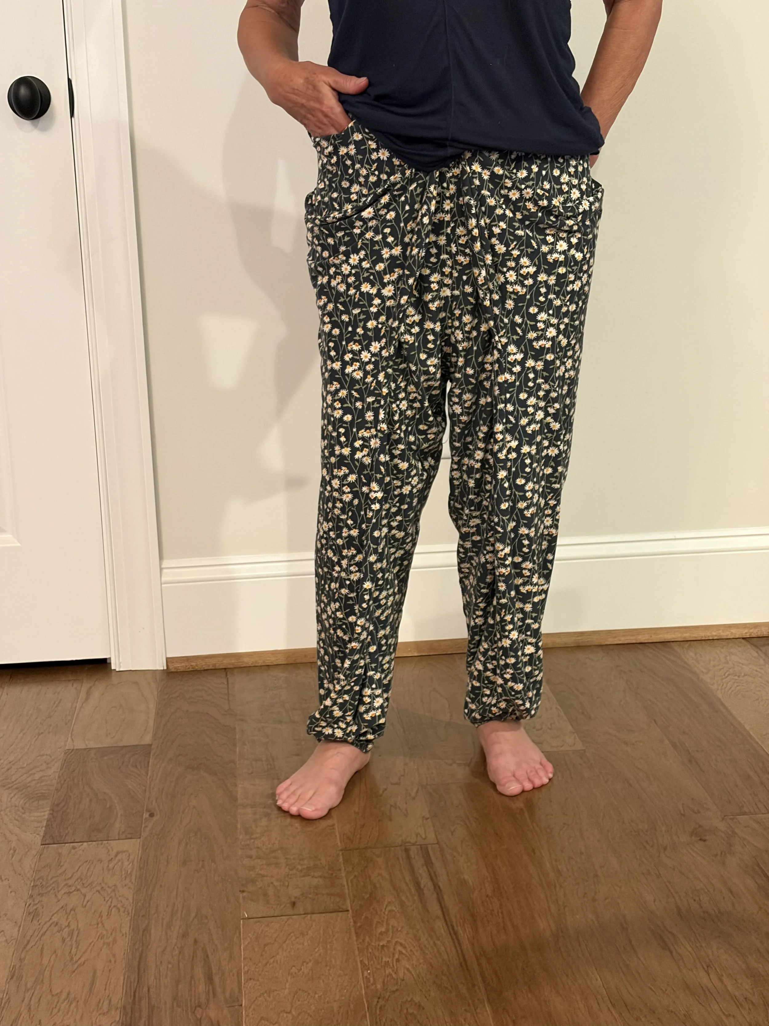 A person wearing black floral lounge pants with a white daisy pattern and a dark navy T-shirt. The person's left hand is in their pocket, and their right hand rests against their hip.