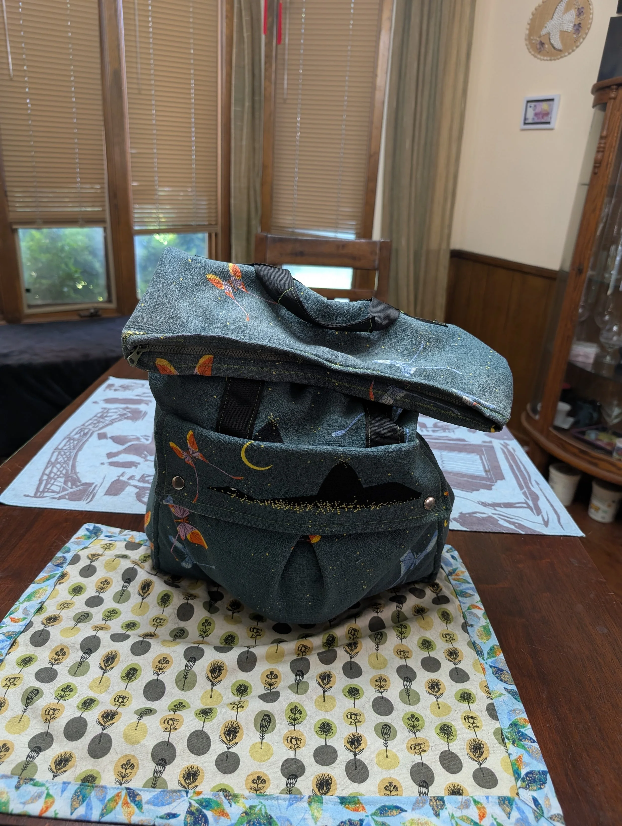 A dark teal fold-over backpack decorated with colorful luna moths, stars, and moons.