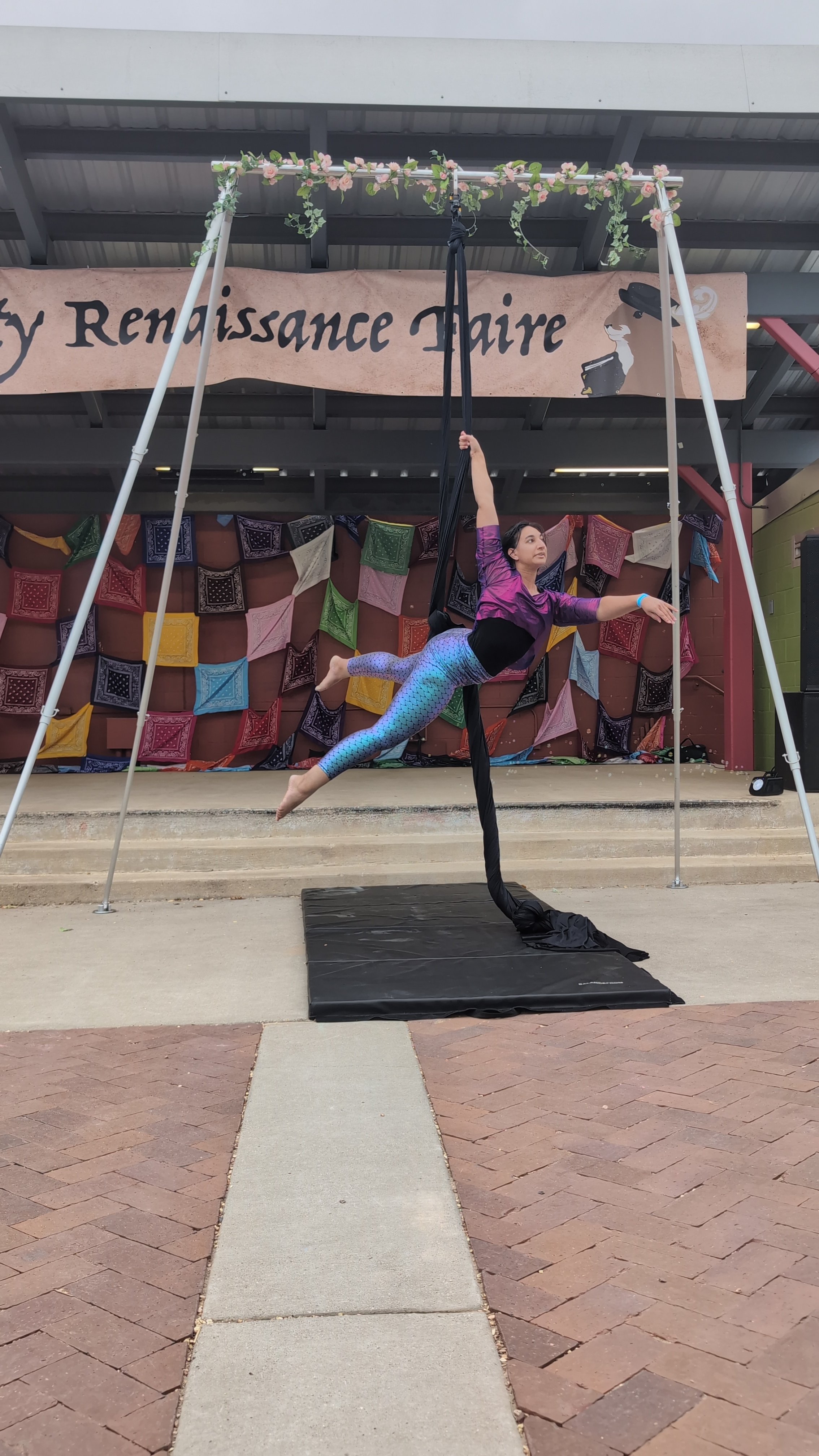 A woman performing an aerial silk routine at an outdoor event. She is hanging horizontally, holding the silk with one hand, with mermaid leggings and a purple top.