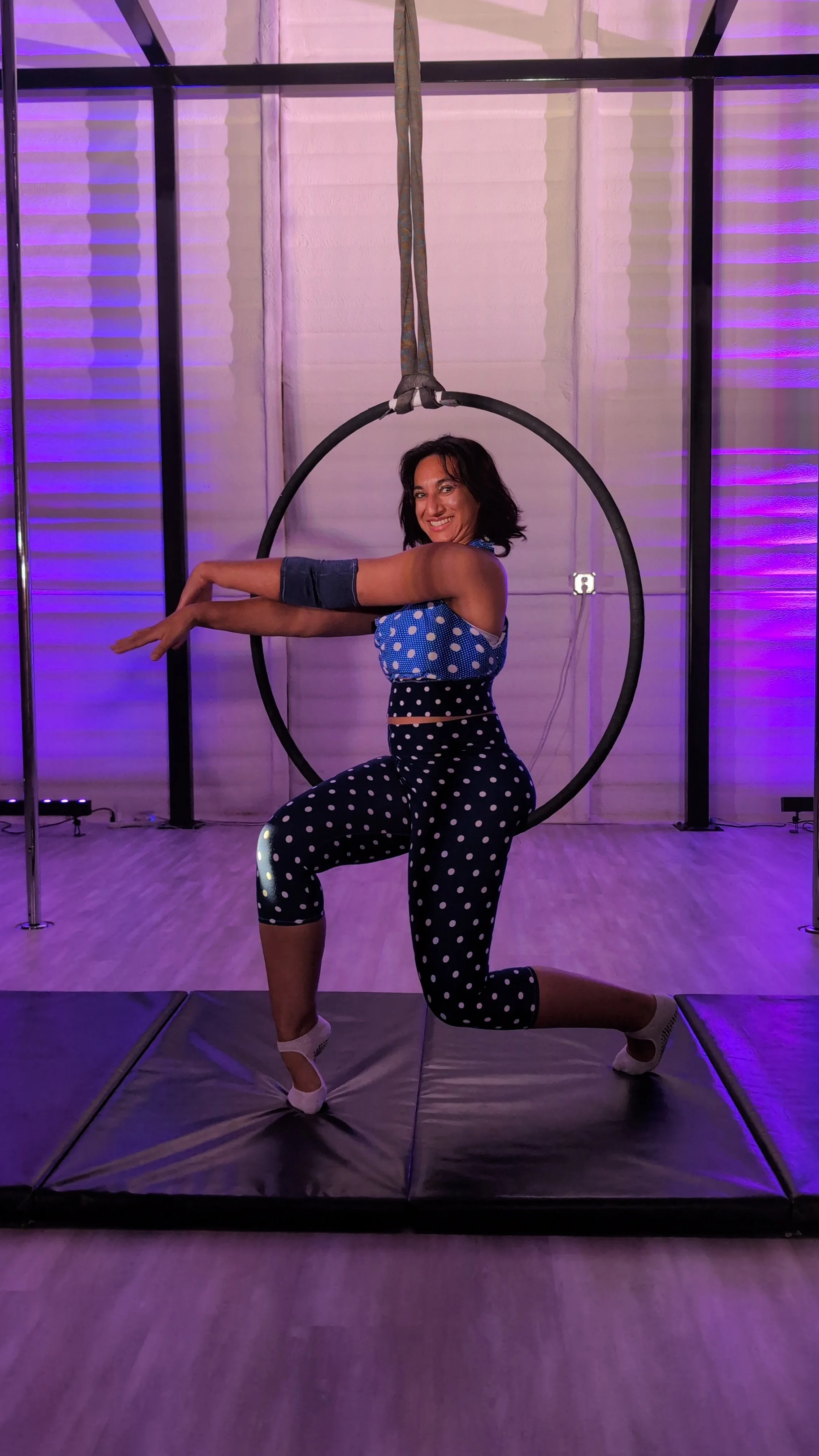 A woman is posing inside a suspended aerial hoop, kneeling on one knee, in a room with purple lighting. She's wearing a blue polka dot top and matching pants with white polka dots, smiling at the camera.