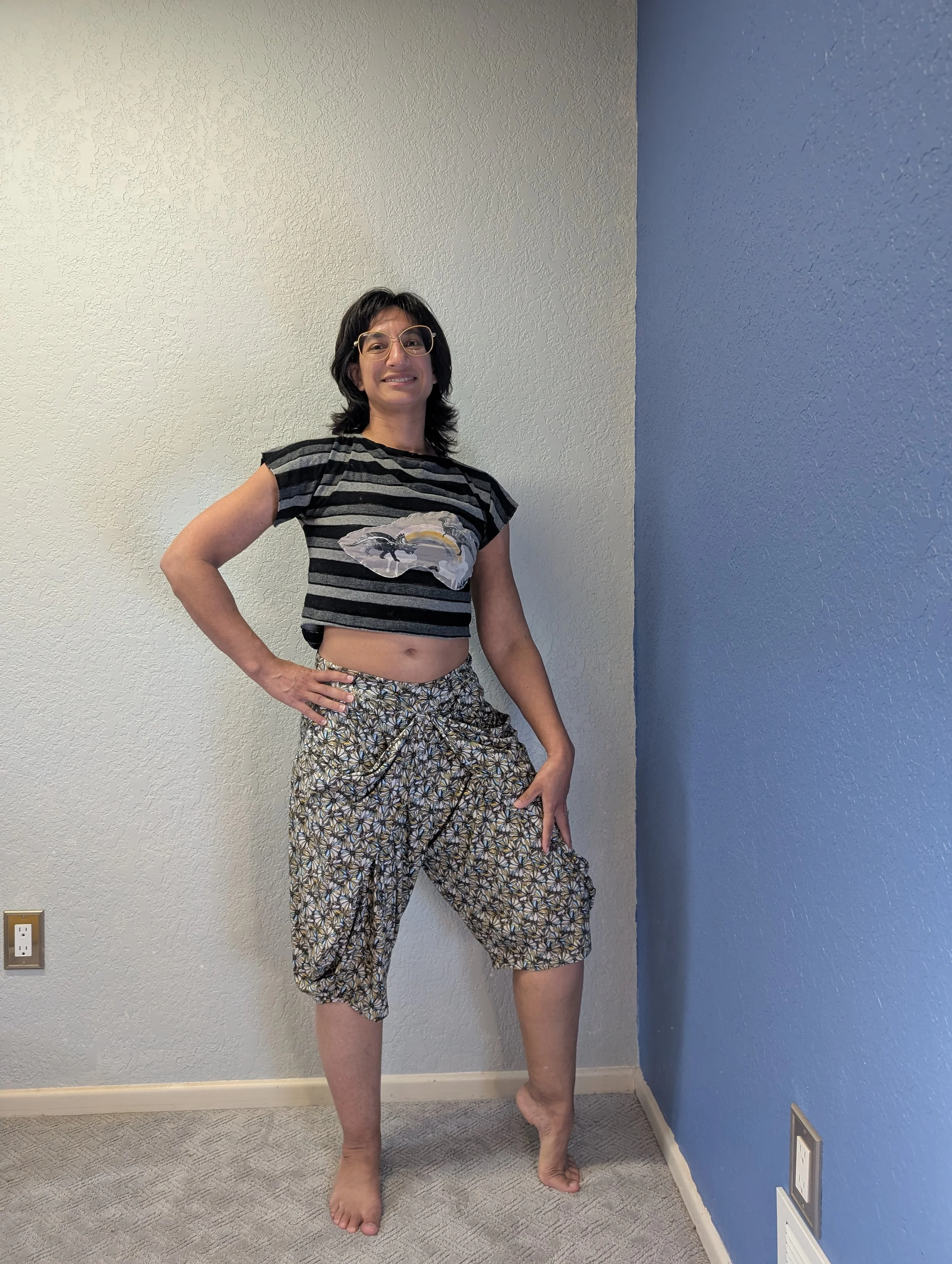 A woman wearing a striped crop top with a dinosaur design and patterned pants with her hand on her hip and her foot on her toes.