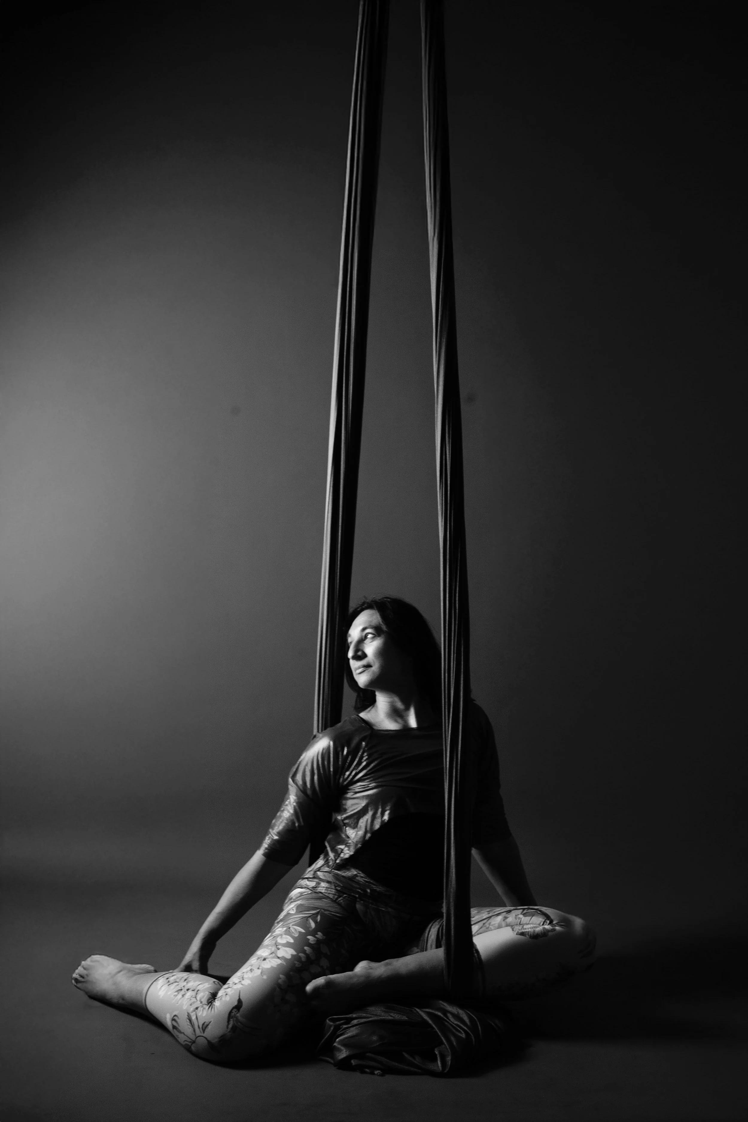 A woman practicing aerial yoga or acrobatics, sitting on the floor with her legs crossed, holding onto aerial silks hanging from above, in a studio with a plain wall background.