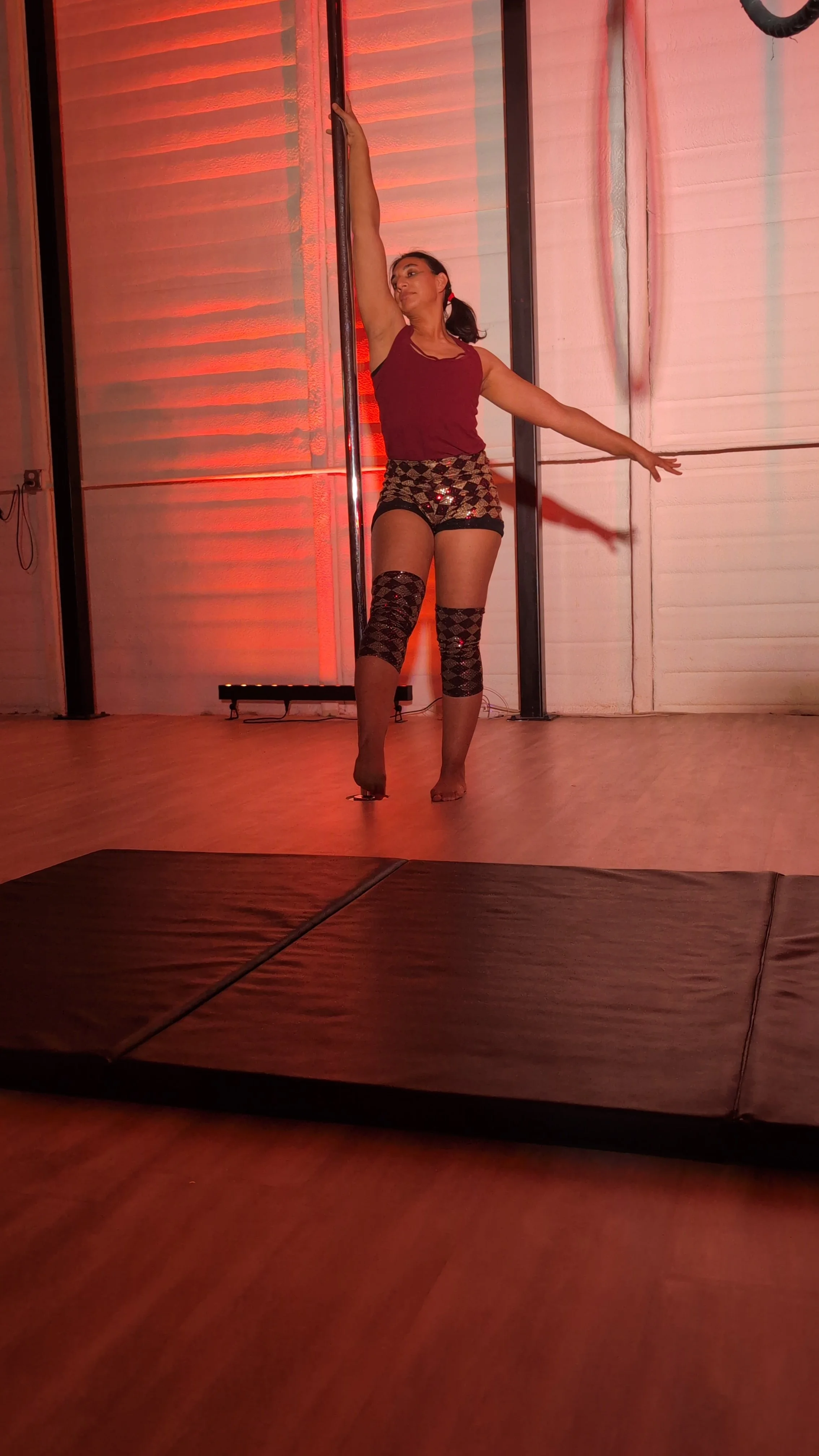 A woman performing a pole dance on a stage with red lighting, wearing patterned shorts, a sleeveless top, and leg warmers.