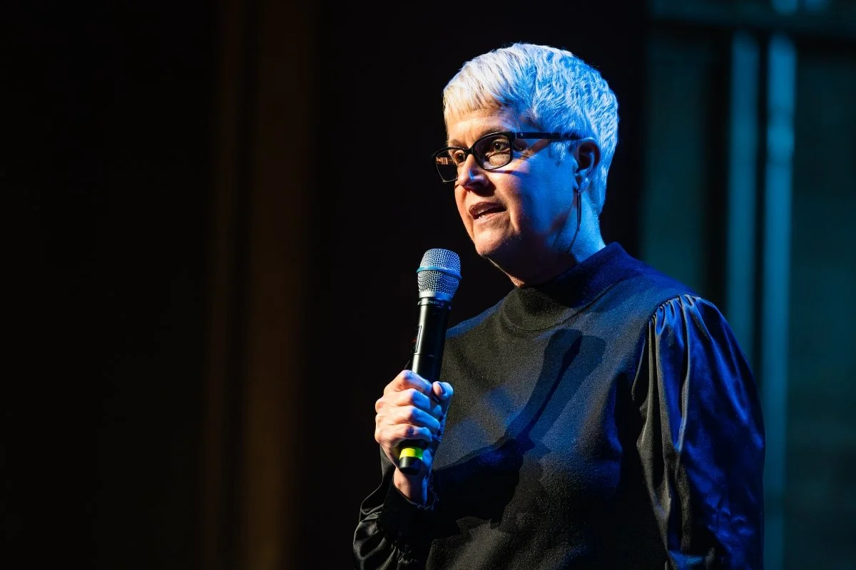 A woman with short, white hair and glasses holding a microphone, speaking during a presentation or performance in a dark setting with stage lighting.