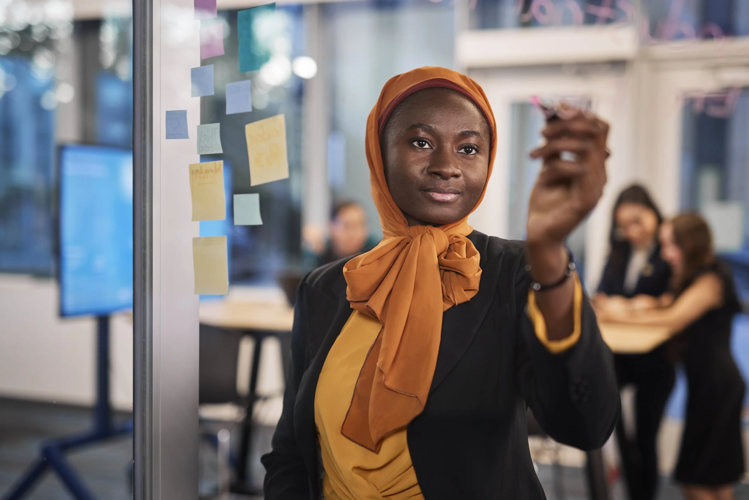 A woman in a black blazer and orange scarf tied around her neck, holding a marker, standing in front of a glass whiteboard with colorful sticky notes, in a modern office setting with other people in the background.