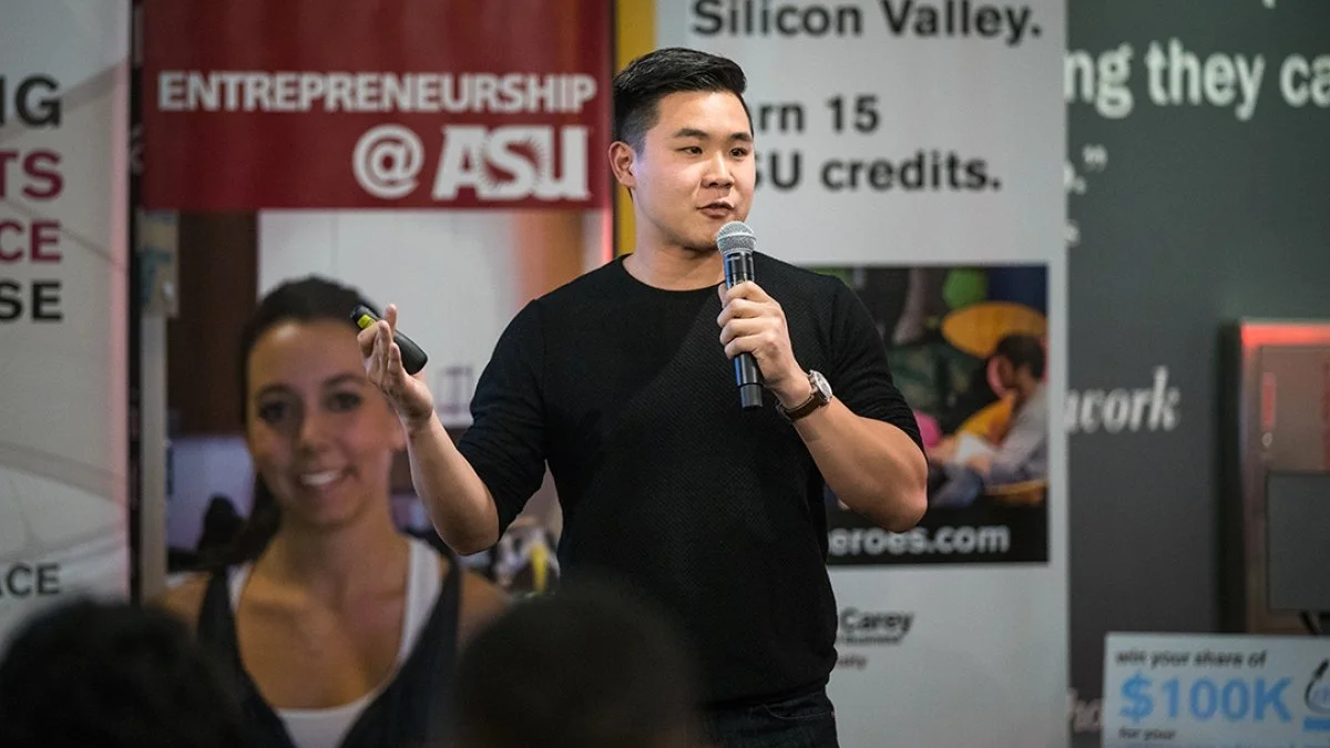 A young man is speaking at an event, holding a microphone and a clicker. A woman is smiling in the foreground. Behind them are banners related to entrepreneurship and ASU.