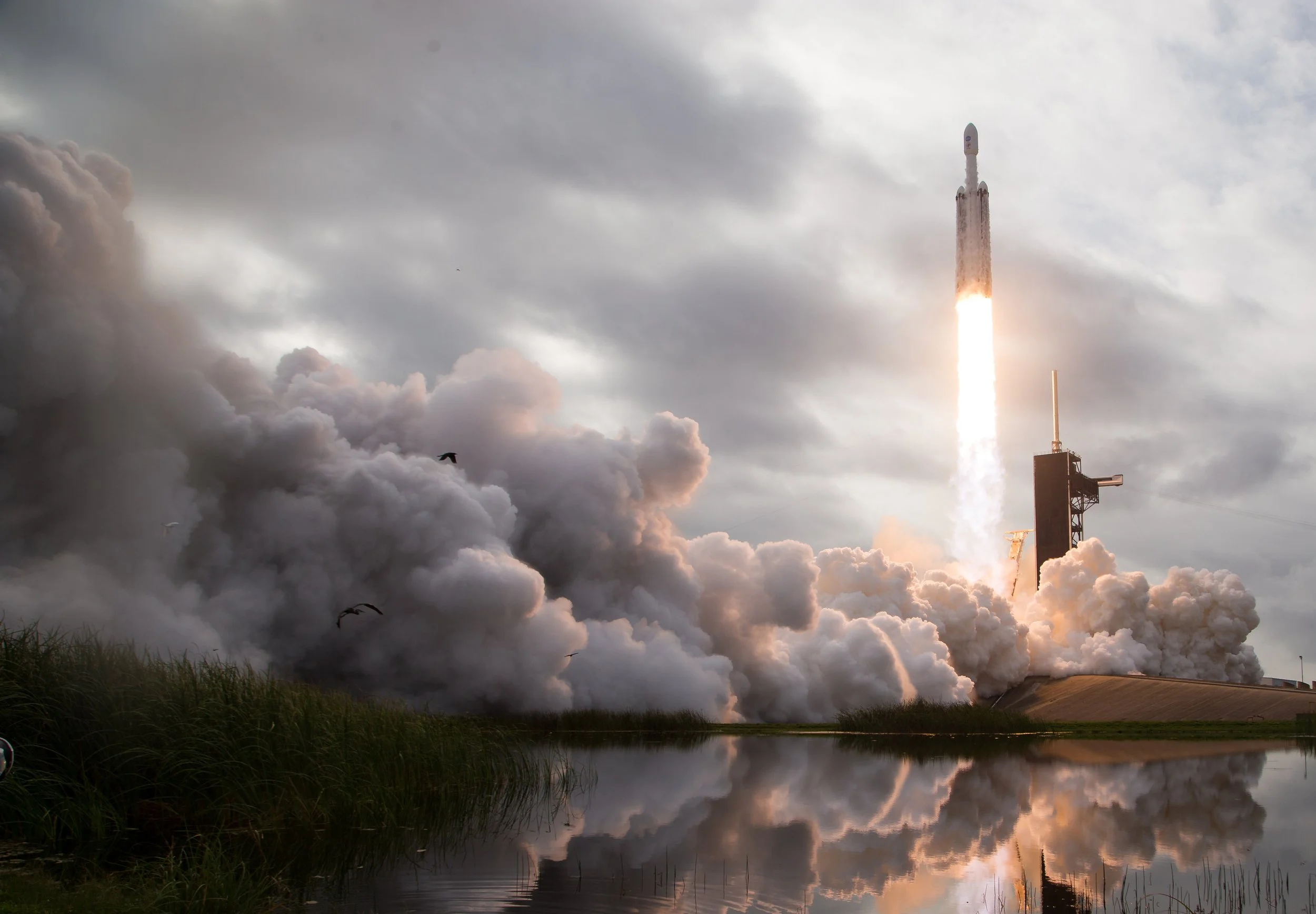 A rocket launches into the sky with smoke and fire, surrounded by dark clouds and reflections on a body of water below.