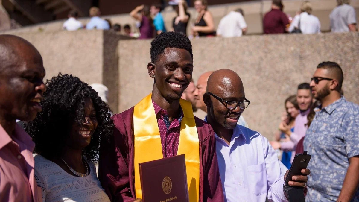 A young man in graduation gown and stole posing with family or friends, celebrating outdoors during a graduation ceremony.