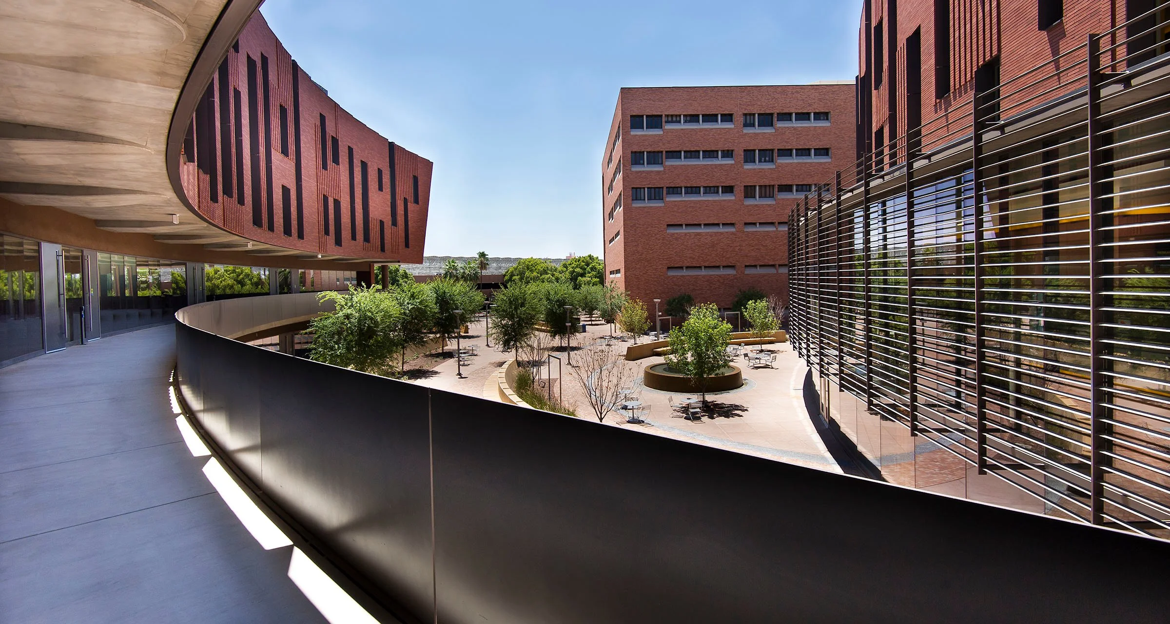 Modern campus courtyard with trees, sitting areas, and multi-story buildings with brick and glass facades.