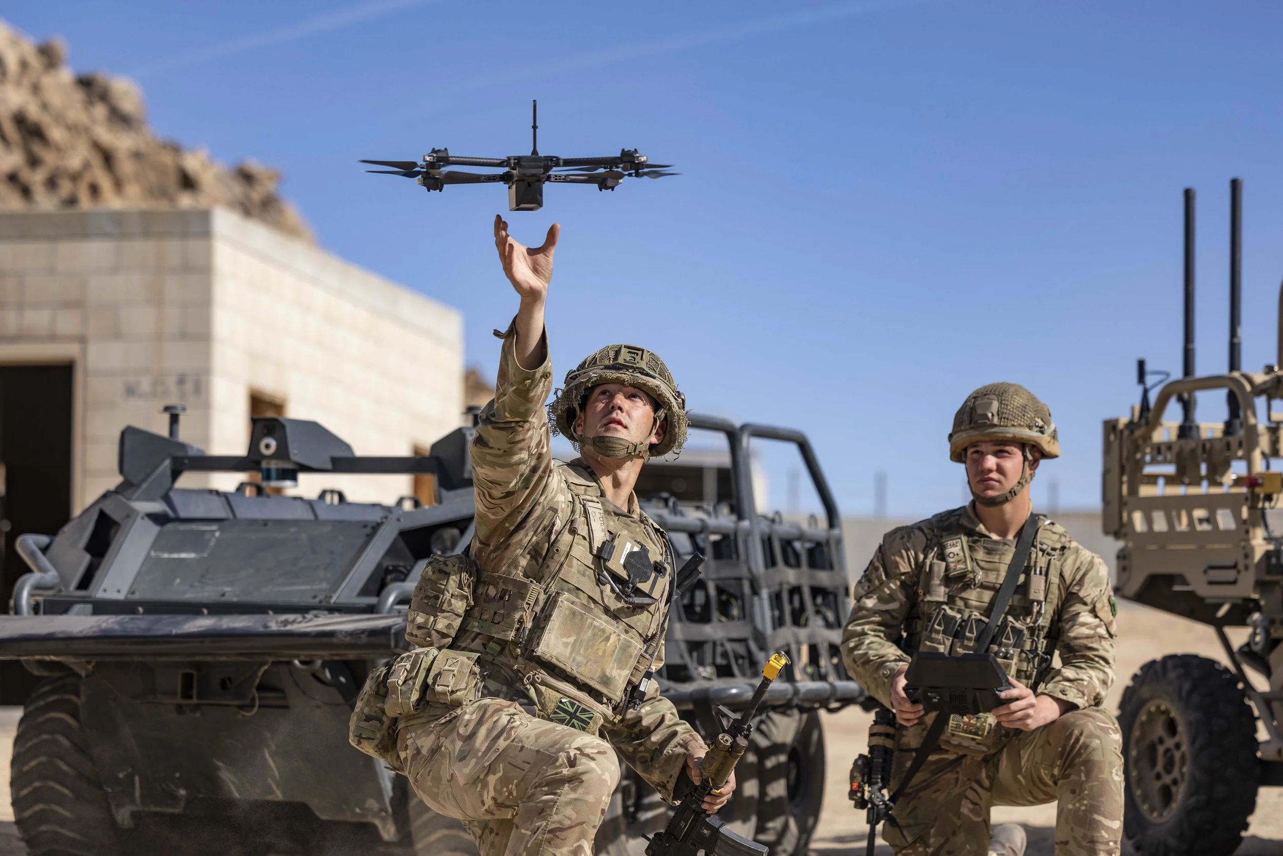 Two soldiers in camouflage uniforms and helmets, one kneeling and the other standing, operating military equipment outdoors under a clear blue sky. The standing soldier is tossing a drone into the air.