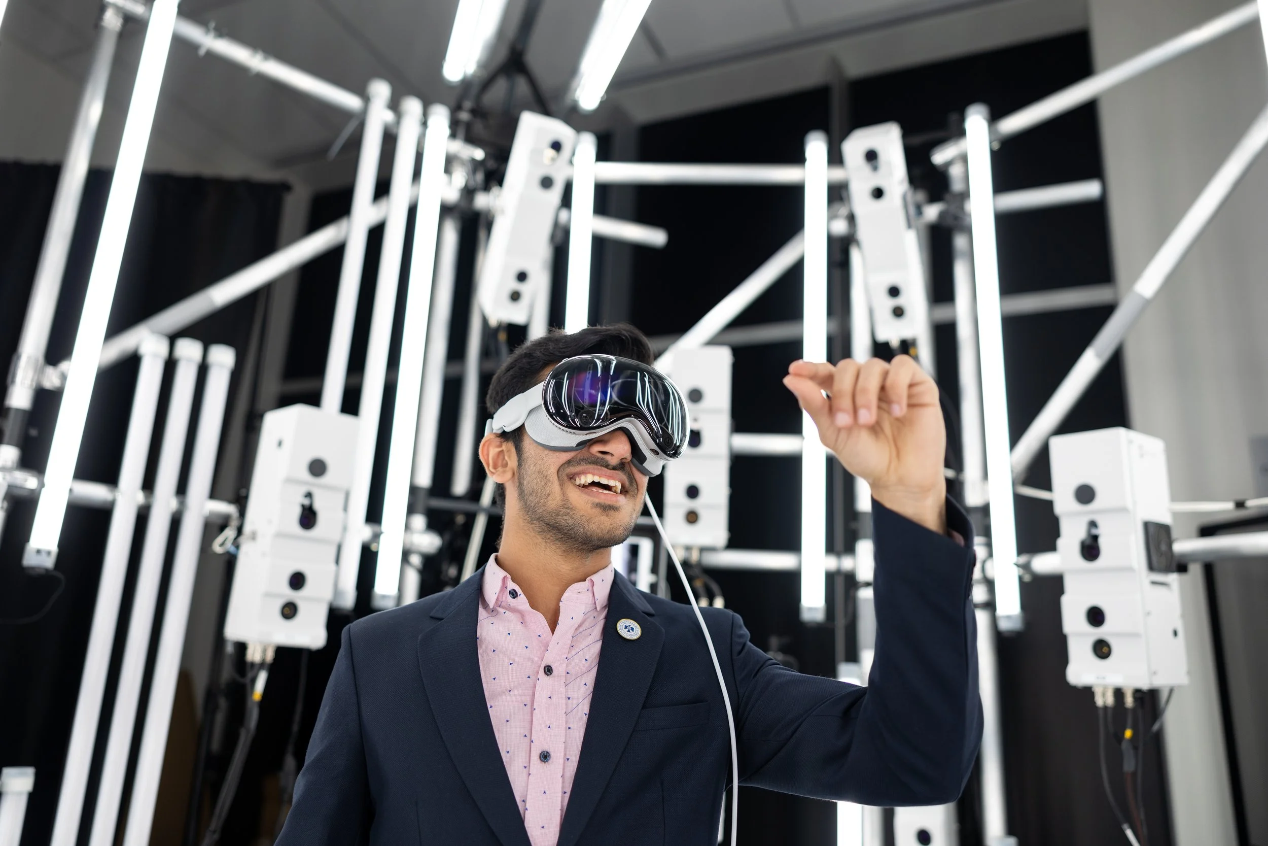 A man in a suit with a pink shirt wearing virtual reality goggles, smiling, and raising his right hand, in a high-tech environment with vertical white lights and machinery.