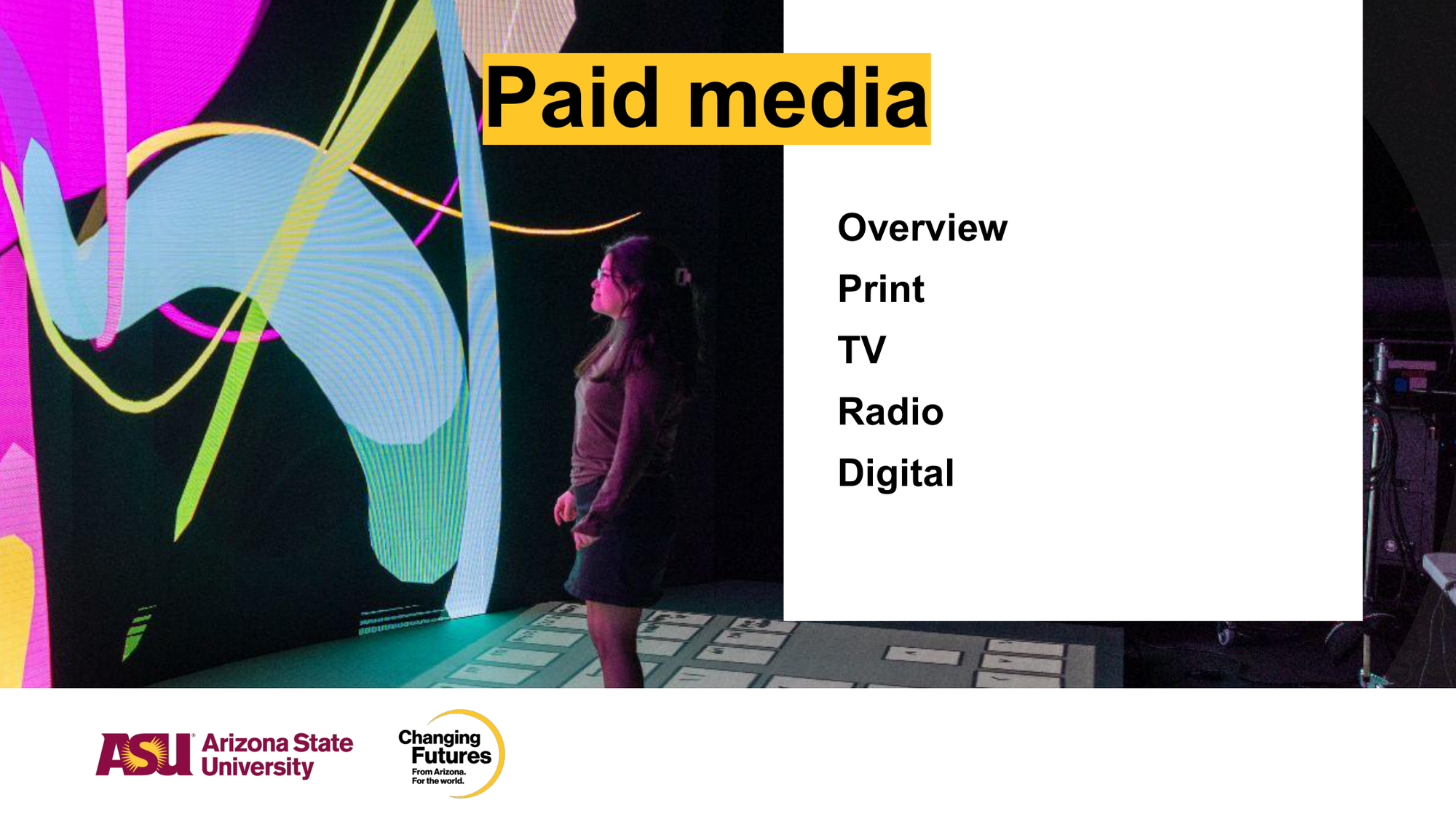 A person standing in front of a colorful digital display at Arizona State University, with a list of categories including 'Overview,' 'Print,' 'TV,' 'Radio,' and 'Digital'.