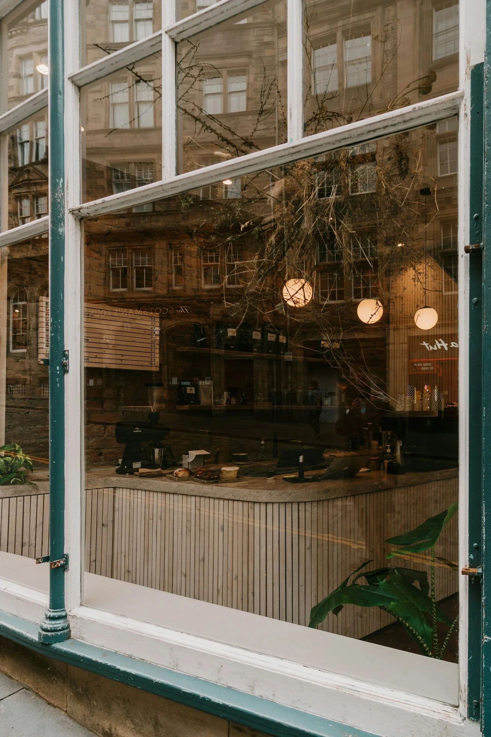 Looking through a large glass window of a bakery or cafe, showing baked goods on display and warm lighting inside. The reflection of nearby buildings and the street can also be seen in the glass.