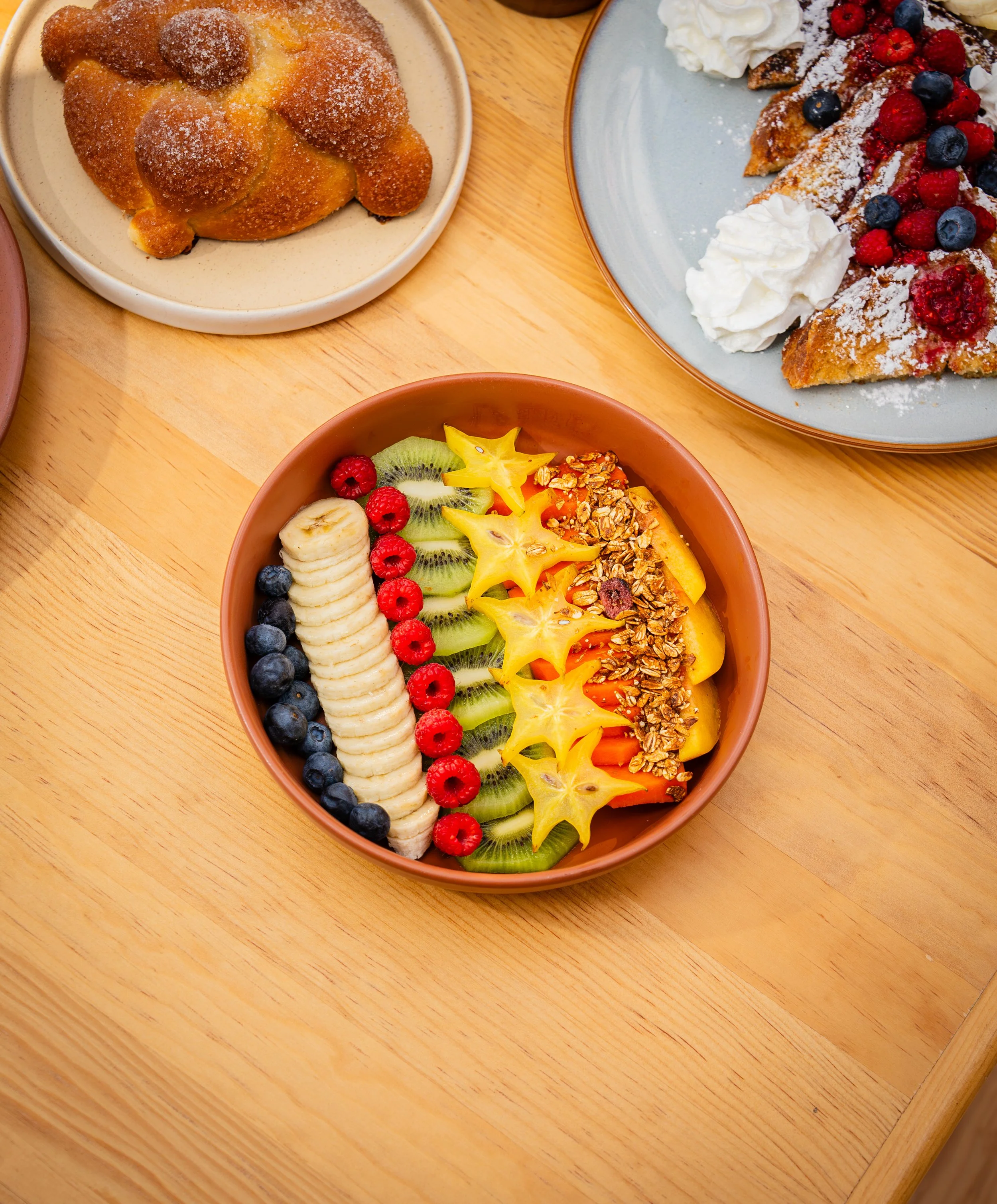Un tazón con frutas picadas, incluyendo plátano, kiwi, frambuesas, arándanos y estrellas de carambola, con granola al centro.