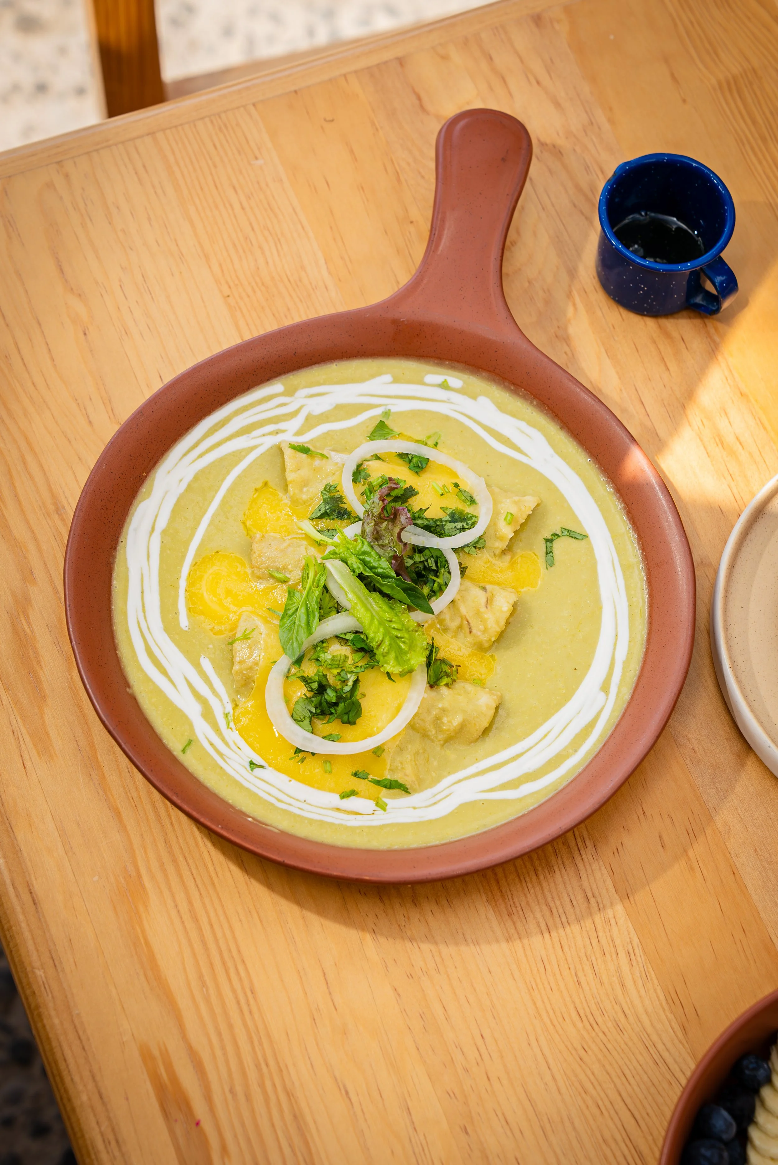 Un plato de comida con crema amarilla, decorada con cilantro, cebolla y trozos de pollo, en un plato de cerámica marrón, acompañado de una taza de café o bebida oscura en un vaso con asa color azul, en una mesa de madera.