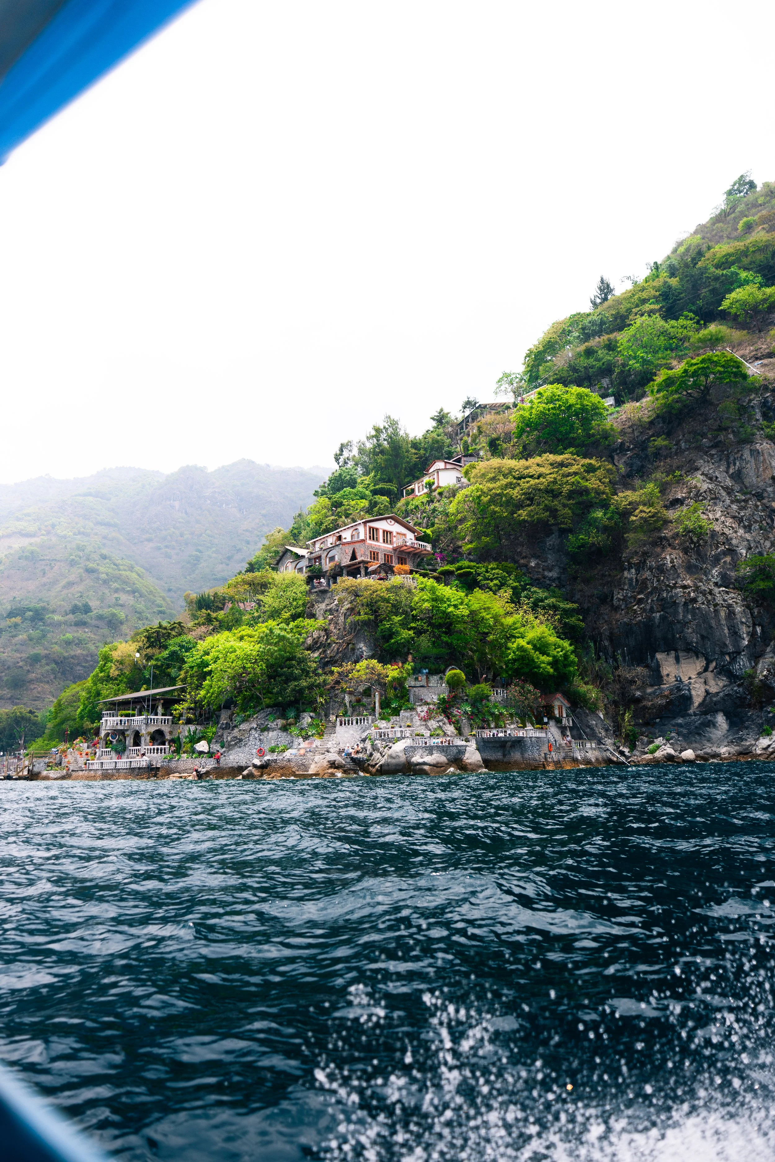 Casas en una ladera de montaña junto a un cuerpo de agua, con vegetación verde y árboles.