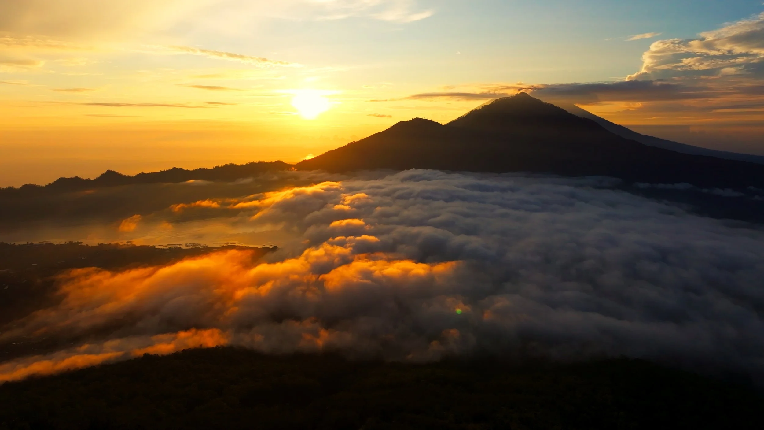 Una vista panorámica del volcán Atitlán rodeado de nubes y con el sol saliendo o poniéndose en el horizonte, creando un cielo con tonos dorados y naranjas.