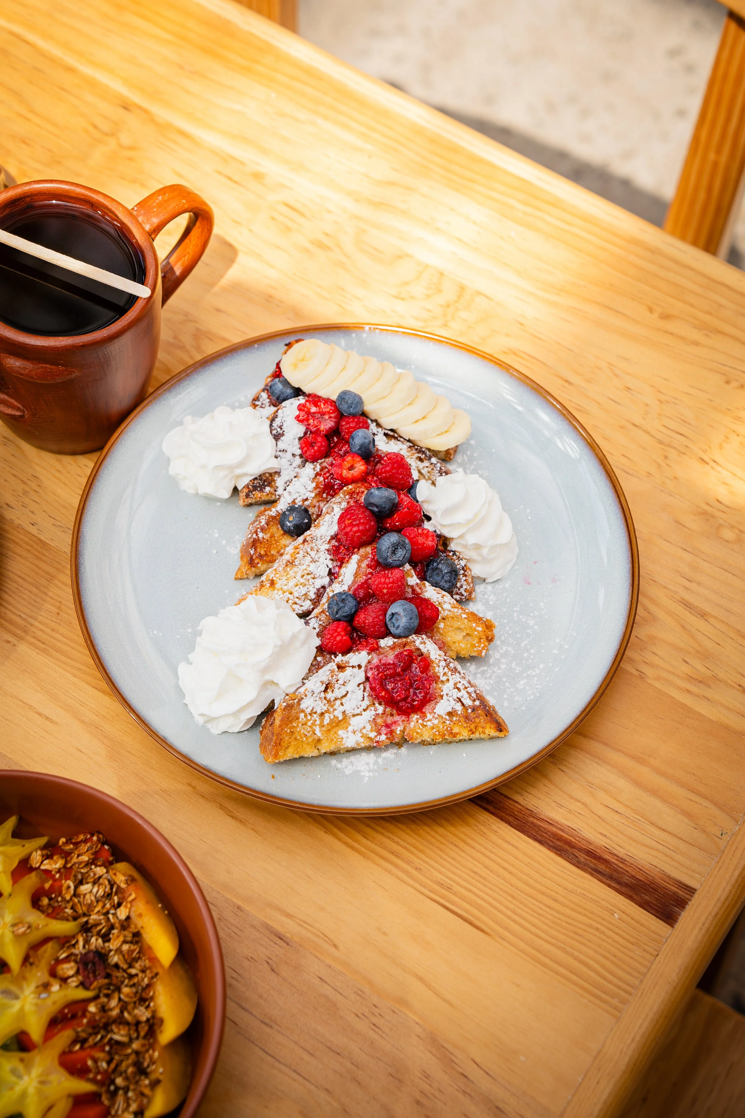 Postre con fruta, crema y polvo de azúcar, acompañado de una taza con una bebida oscura y un palillo