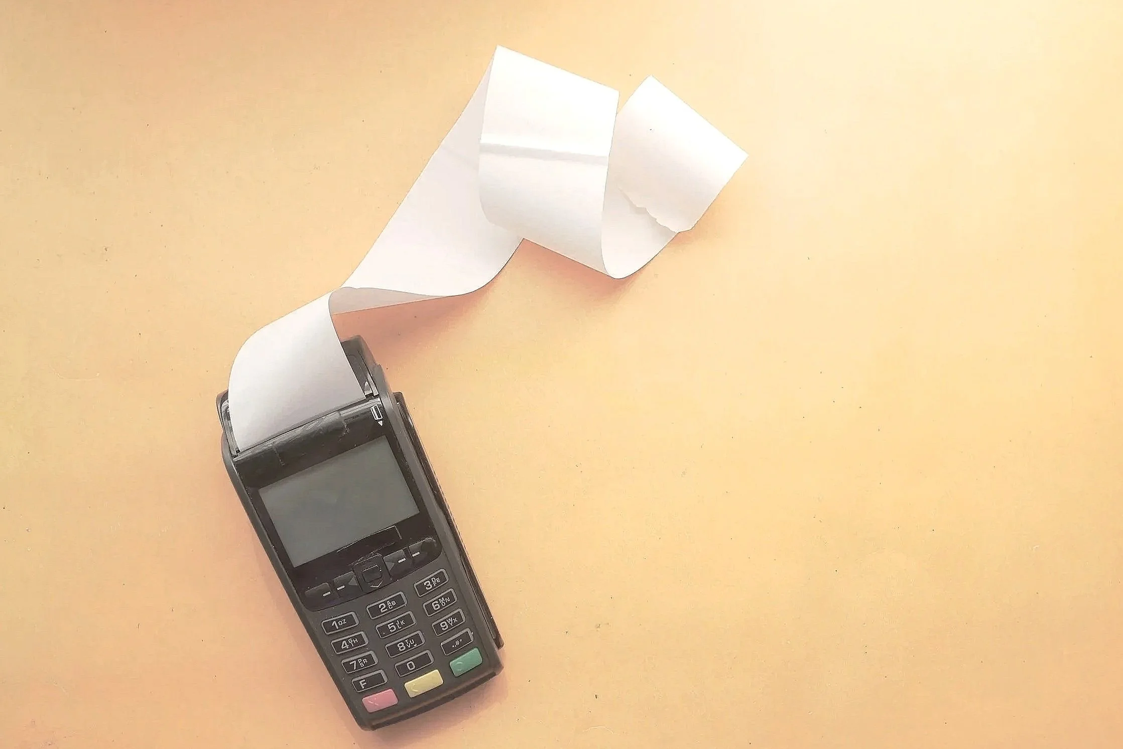 A black electronic payment terminal with a small screen and keypad, next to a white receipt on a pale yellow surface.
