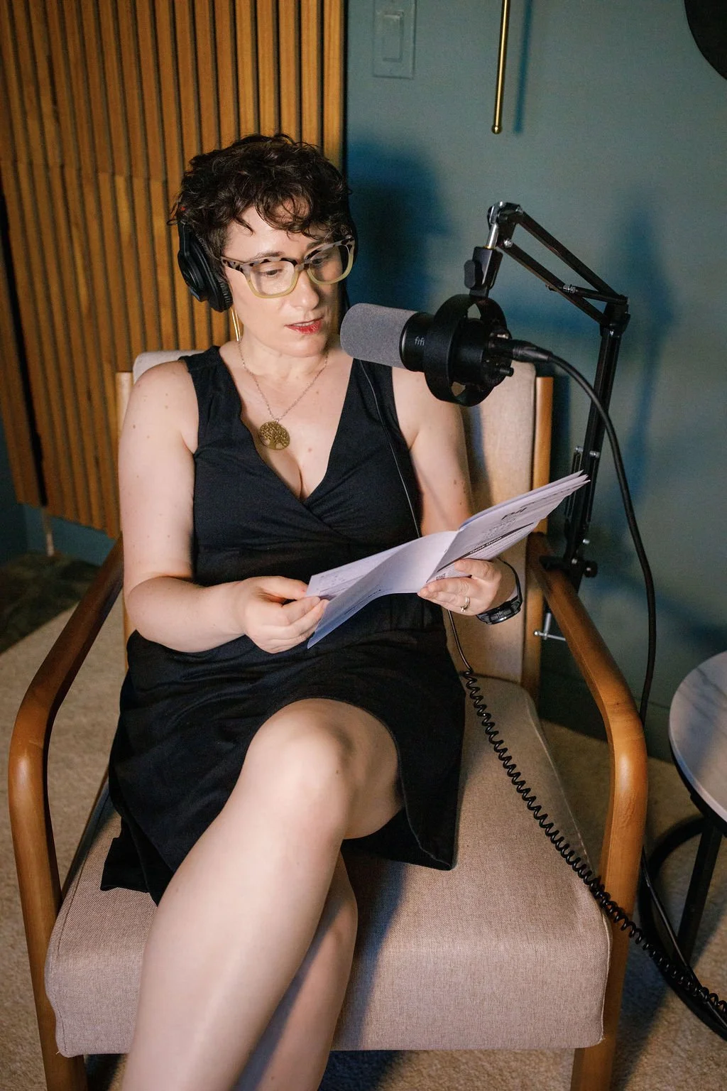 A woman with short curly hair, glasses, and wearing a black sleeveless dress, seated in a chair with a wooden frame, reading a script or sheet music in front of a microphone. She is in a recording studio wearing headphones.