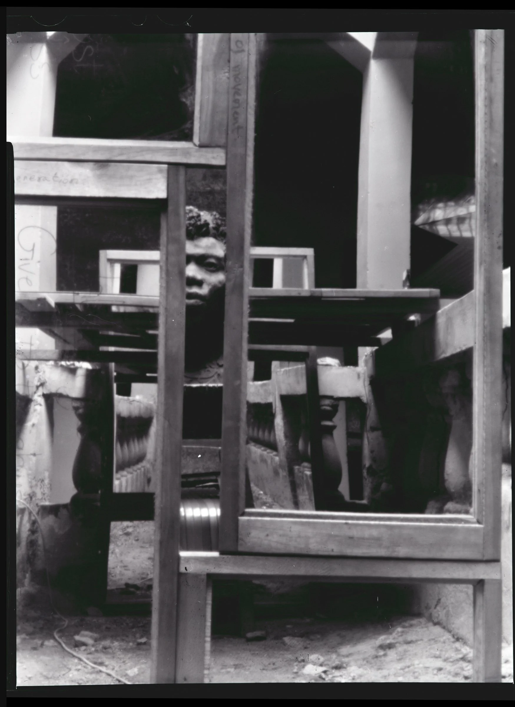 Black-and-white photo of a bust sculpture peeking through the wooden frames of multiple mirrors arranged around it. There are architectural structures, including concrete pillars and a series of concrete balusters, reflected in the mirrors.