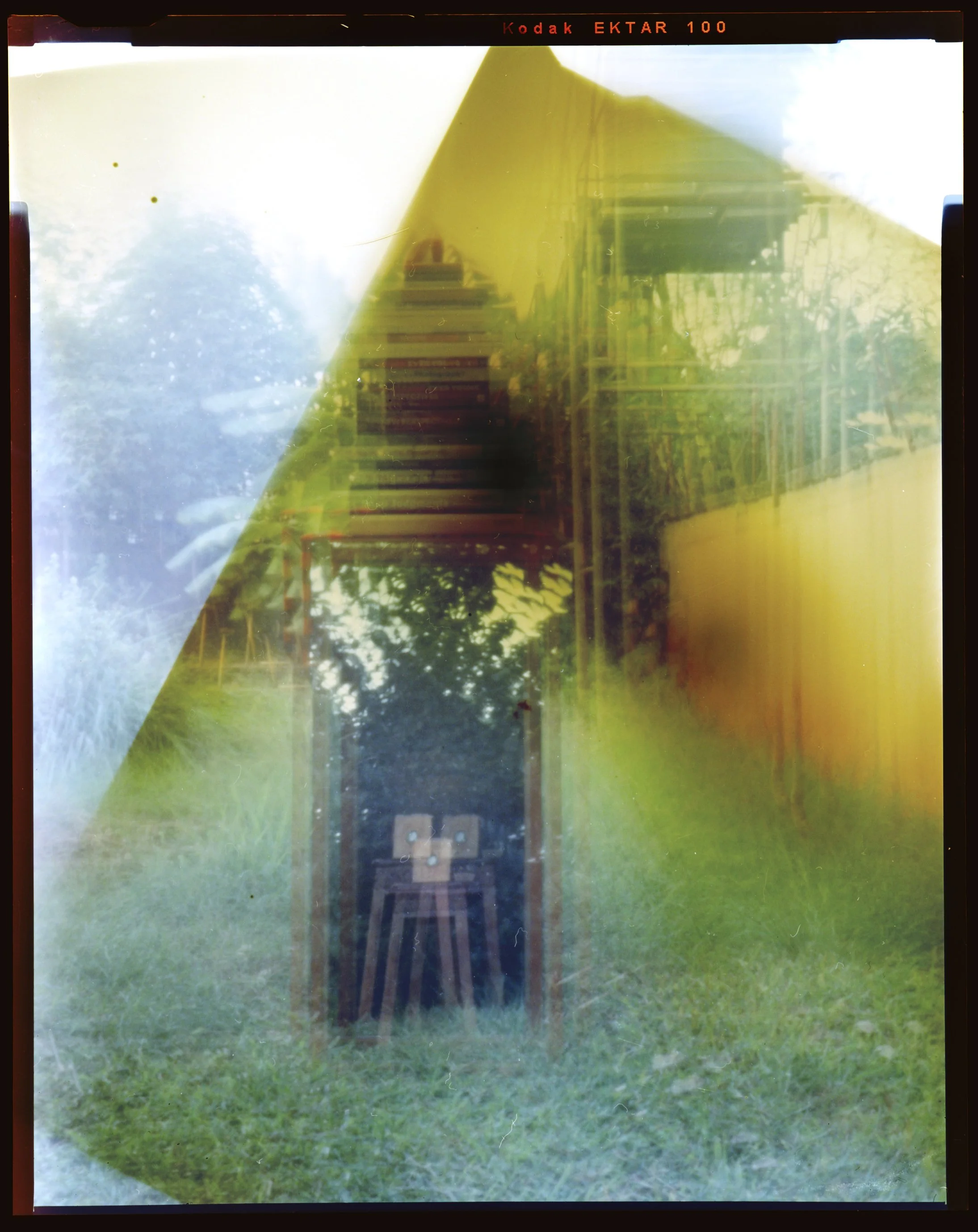 A single-exposure photograph made with a pinhole camera that has 3 holes. It depicts a grassy outdoor area with a mirror propped against a stool with a stack of books. The image appears to be superimposed 3 times because of the multiple pinholes.