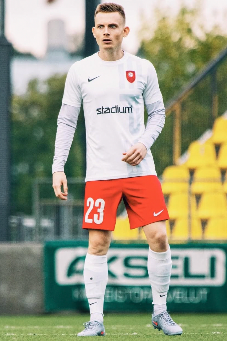 A male soccer player standing on a field, wearing a white jersey with red shorts, number 23, and gray cleats.