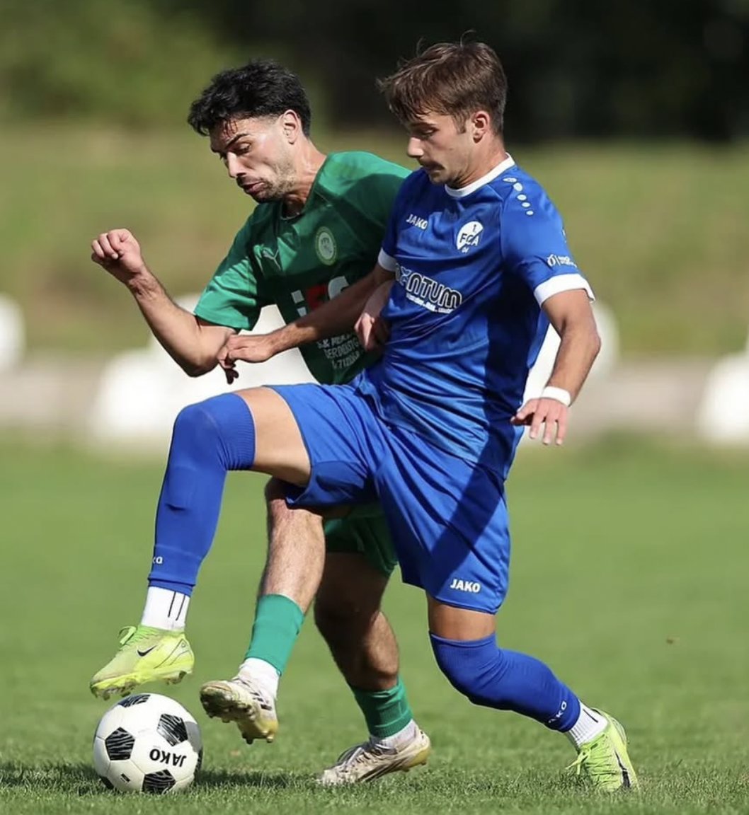 Two soccer players in green and blue uniforms battling for the ball on a grassy field during a match.