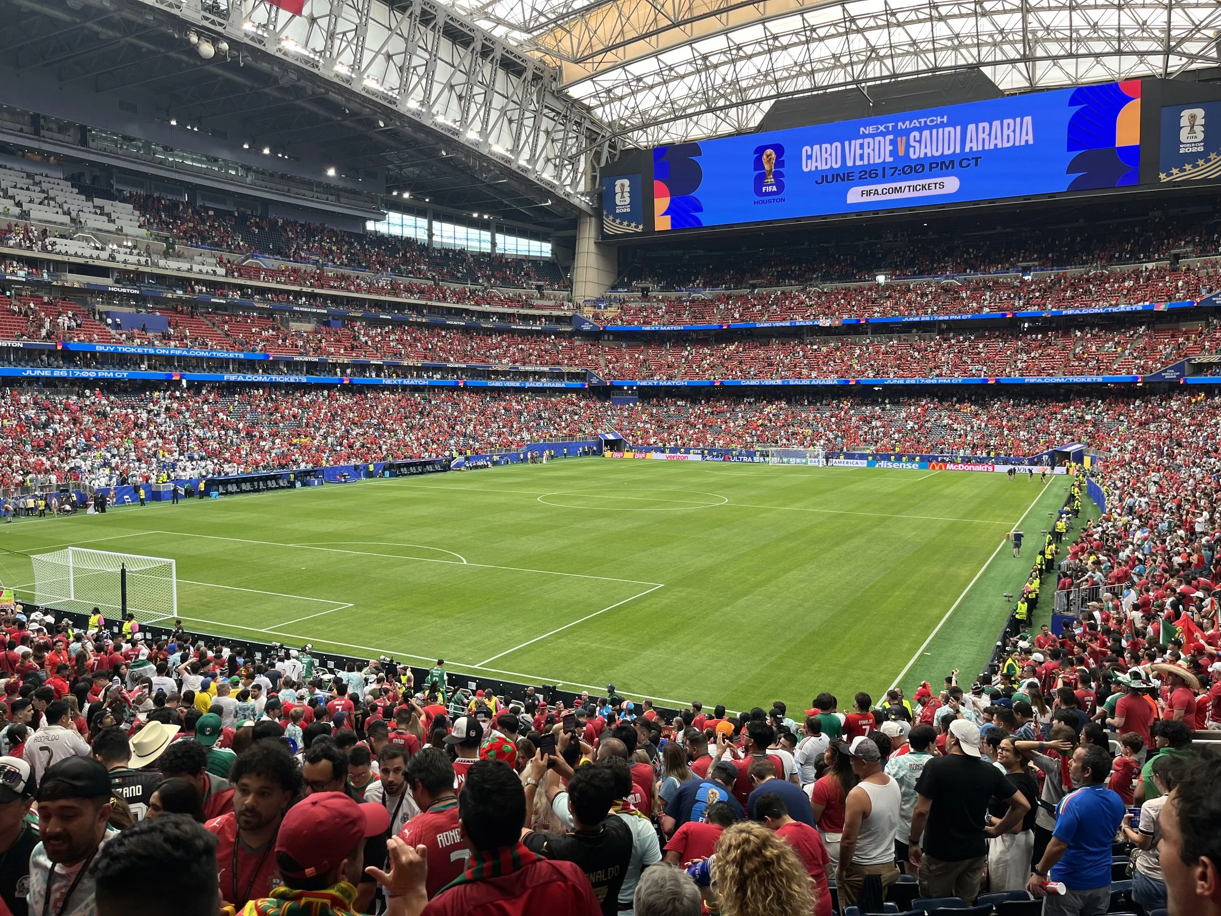 Inside a packed football stadium with a green playing field, cheering spectators in red shirts, and large electronic screens displaying information about an upcoming match between Cabo Verde and Saudi Arabia, scheduled for June 26 at 7:00 PM CT.