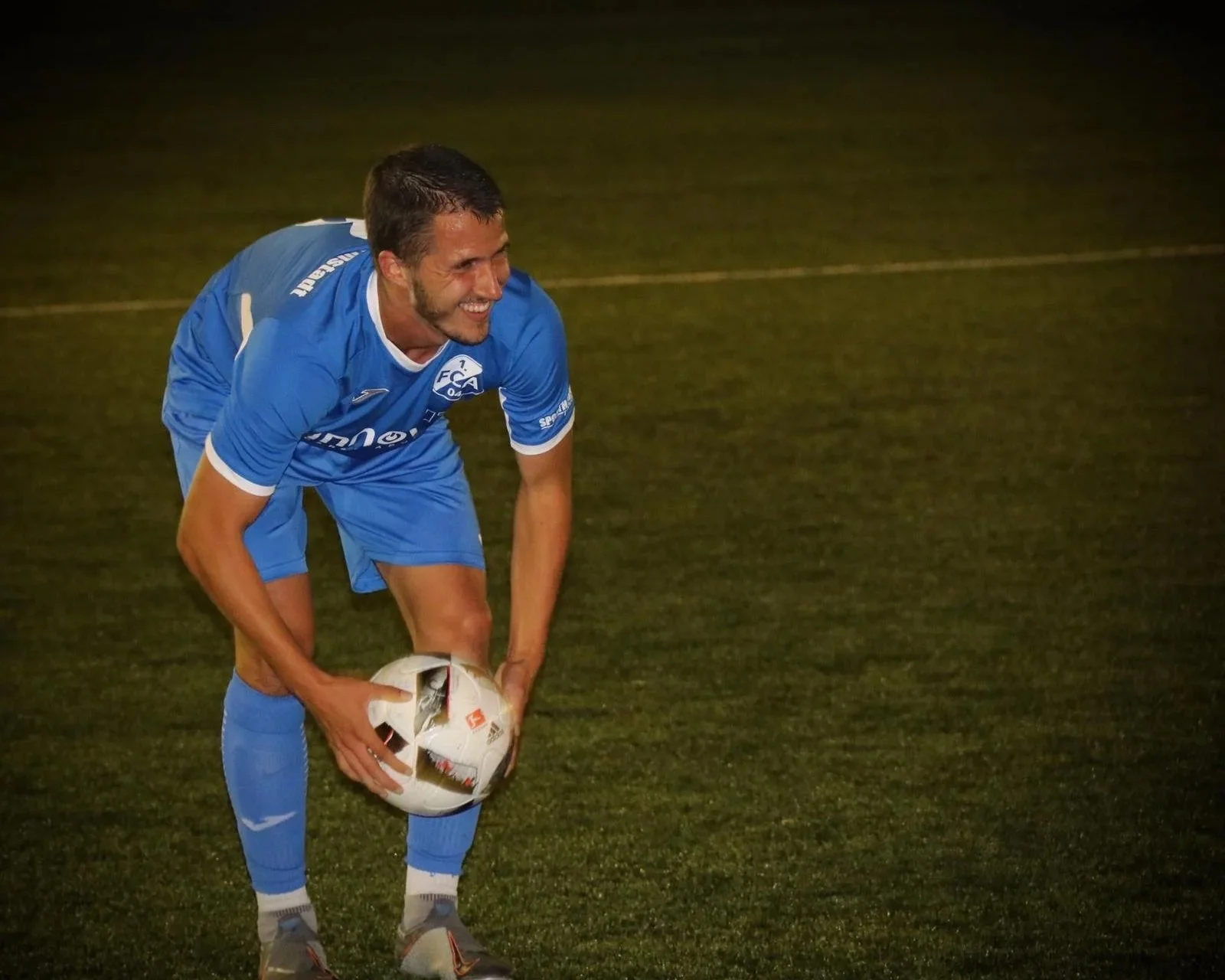 A male soccer player in a blue uniform crouching on a grass field, holding a soccer ball with both hands, smiling, during a night game.