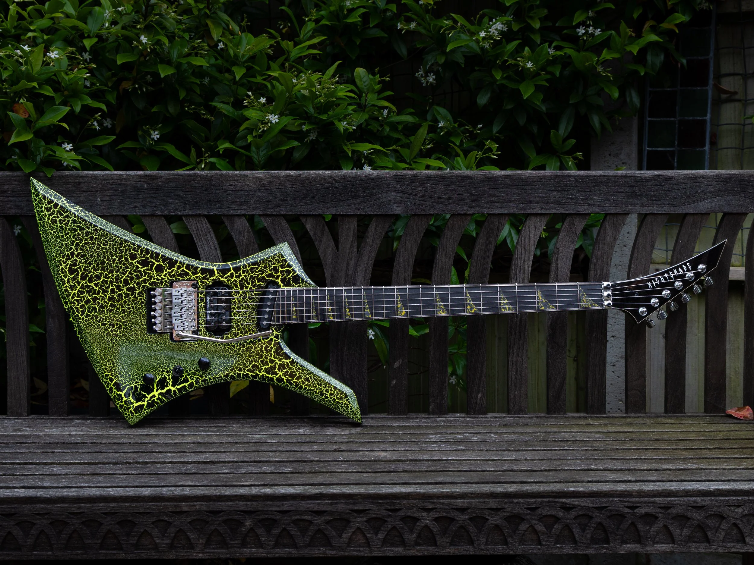 Close-up of a yellow and black electric guitar with a cracked paint pattern, leaning against a weathered wooden fence with green leafy bushes in the background.