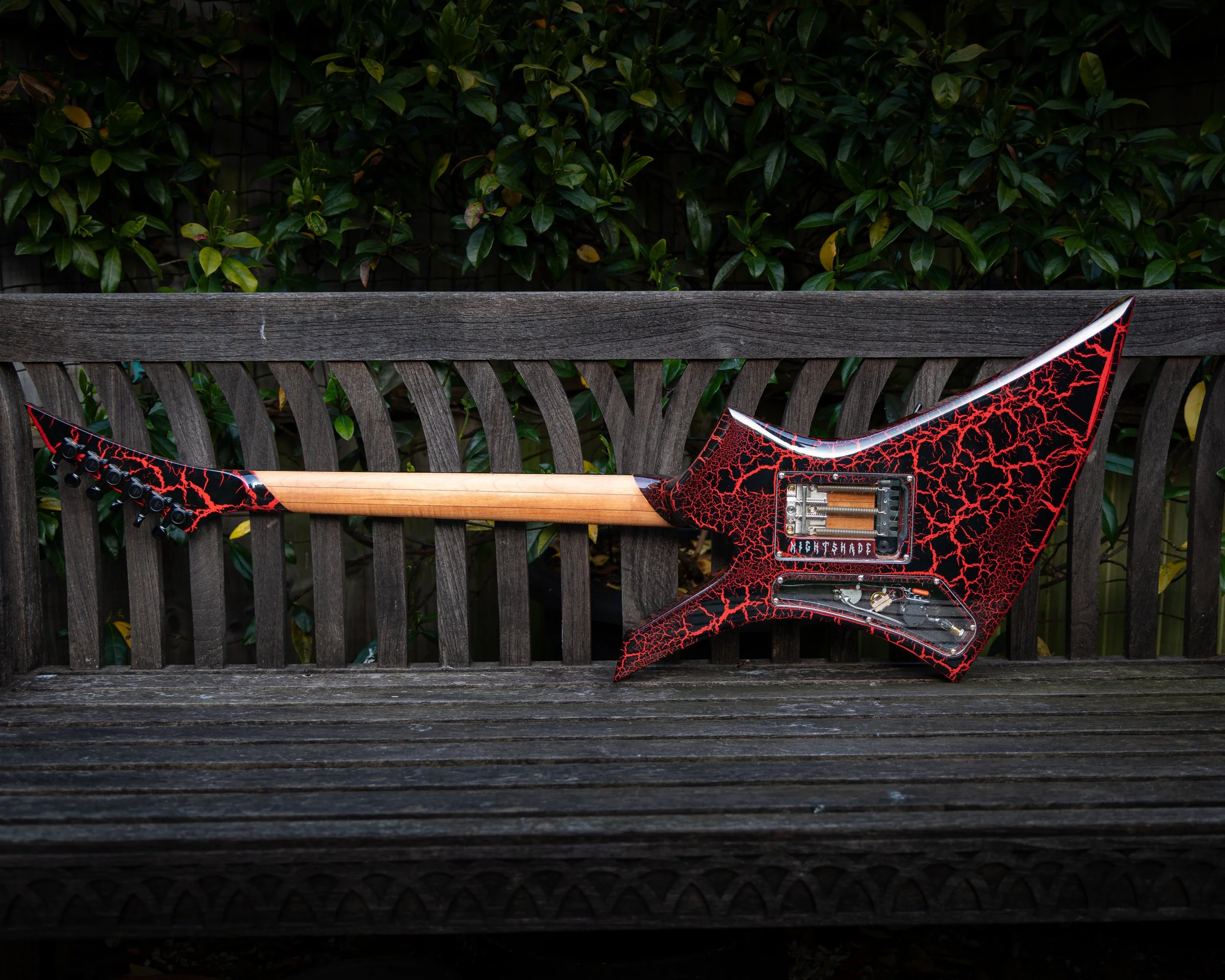 Part of an electric guitar with a red and black cracked pattern design is hanging on a wooden bench in front of green leafy bushes.