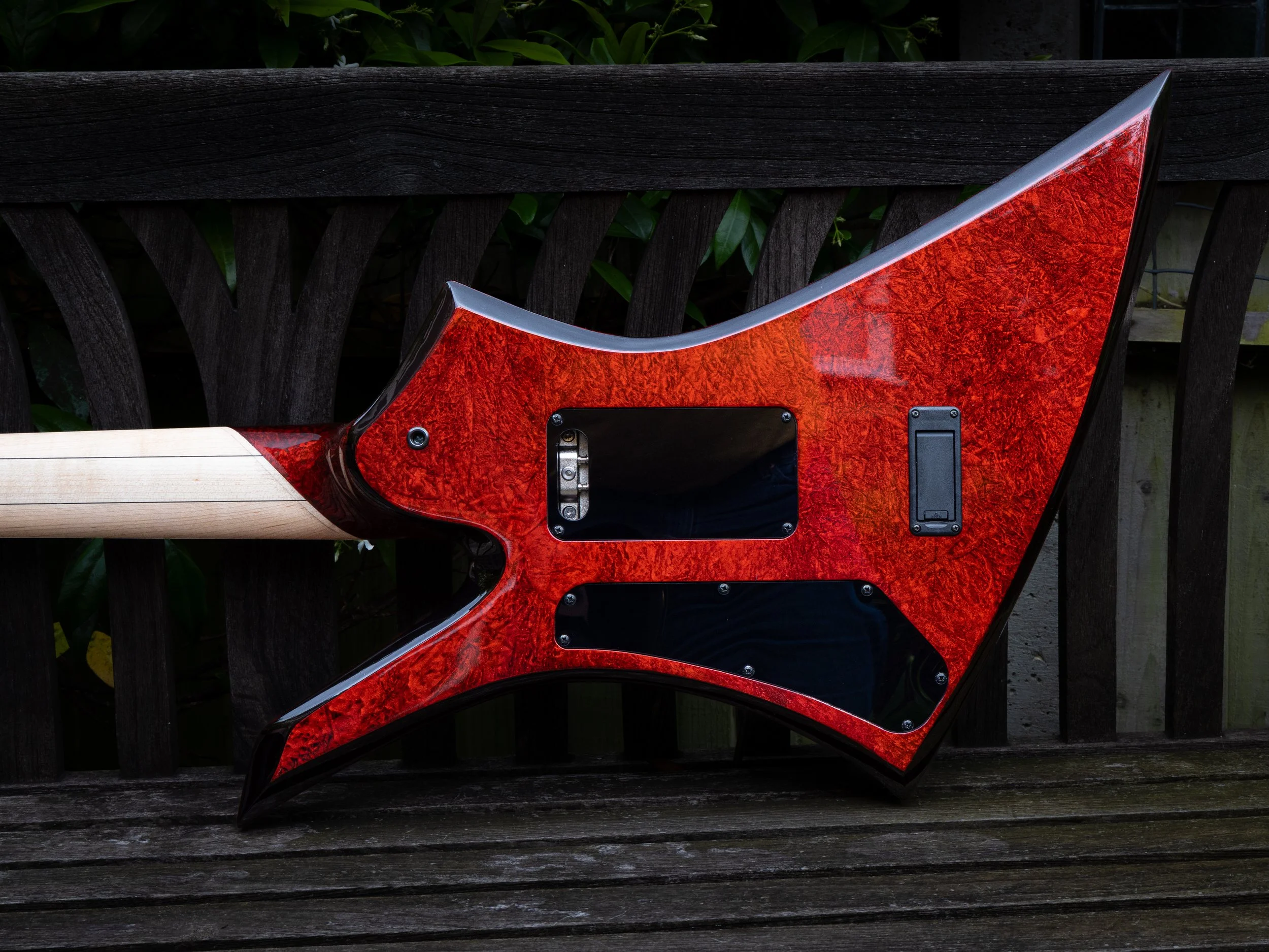 Close-up of a red electric guitar body with a black pickguard, resting on a wooden surface with dark outdoor foliage in the background.
