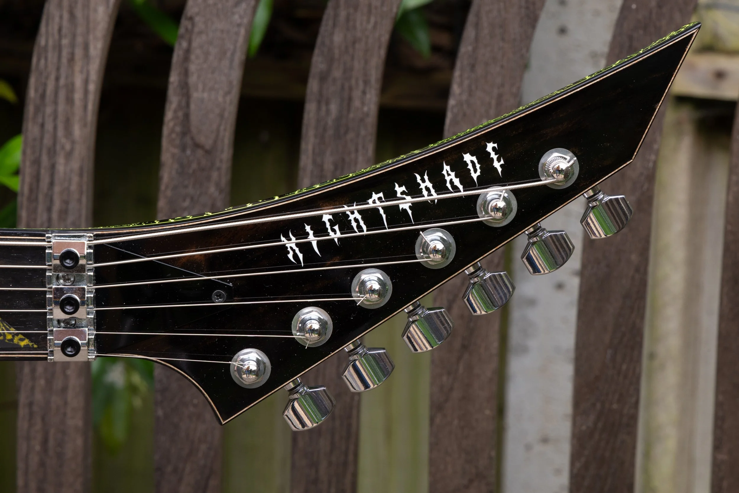 Close-up of an electric guitar's headstock showing tuning pegs, strings, and part of the guitar's logo, with a wooden fence and green foliage in the background.