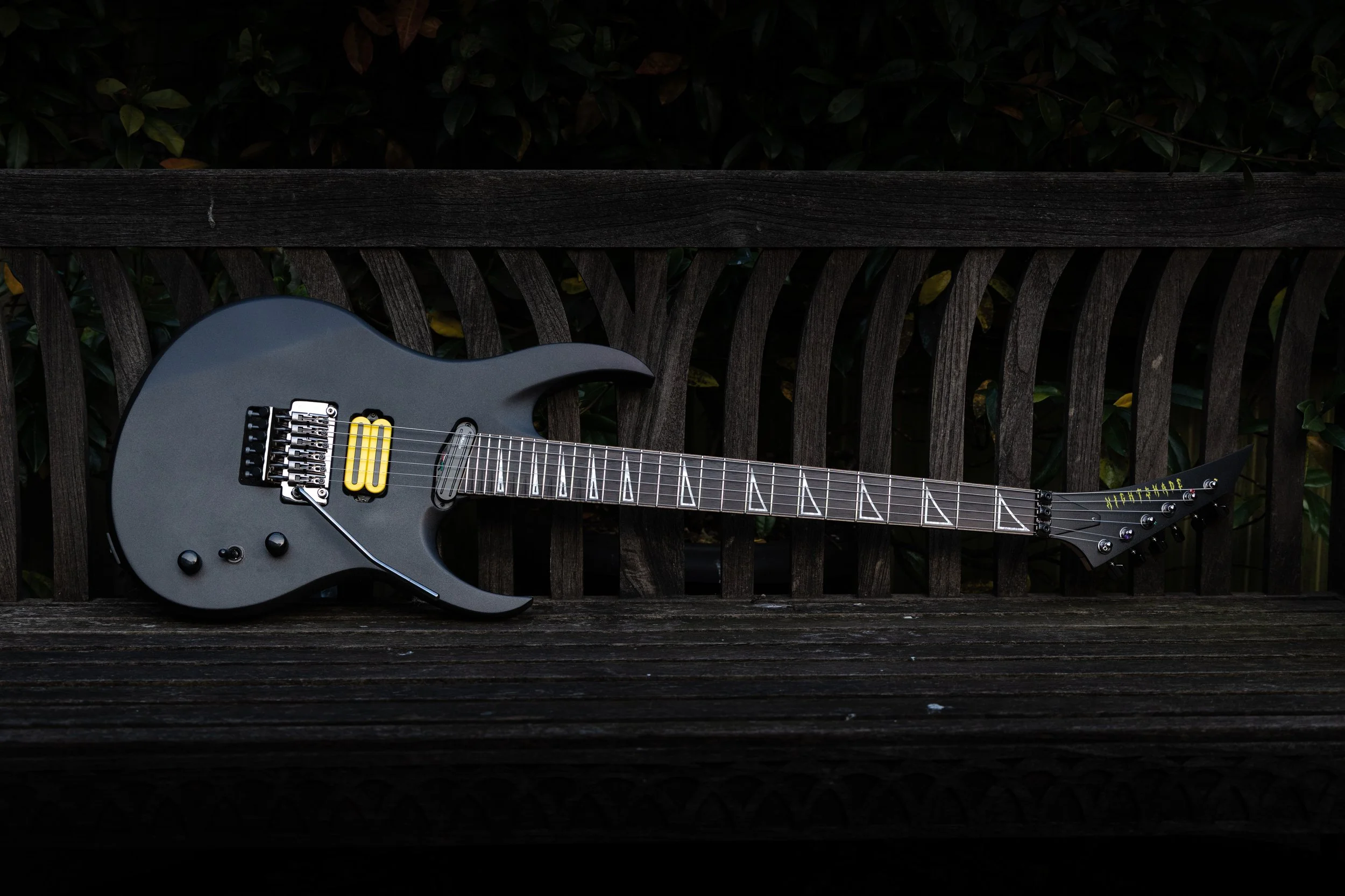 A black electric guitar with yellow pickups leaning against a dark wooden bench outdoors, with foliage in the background.